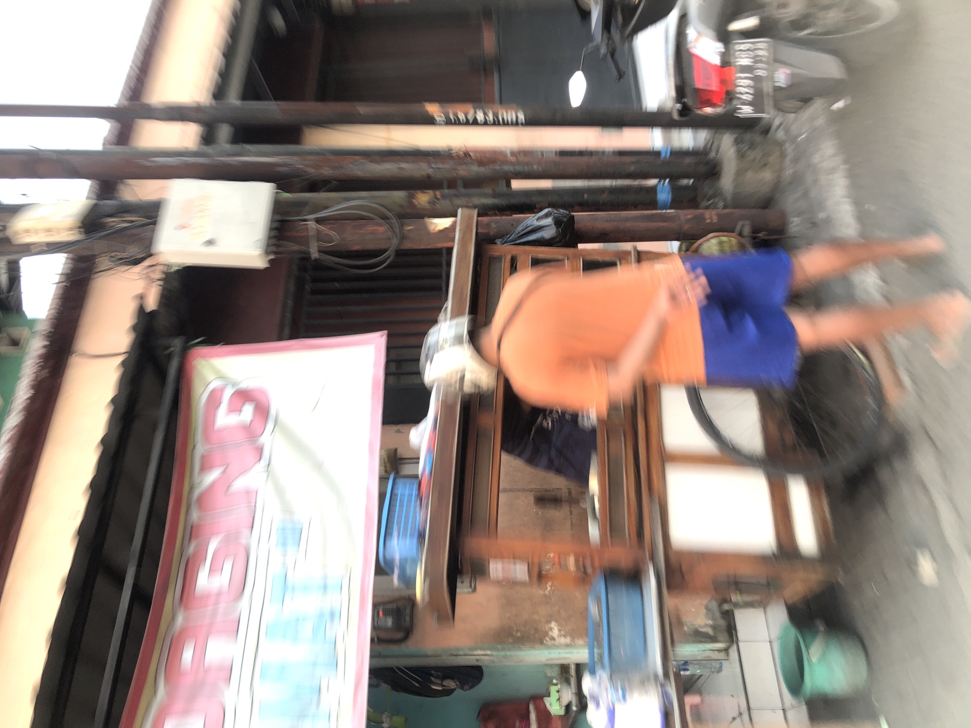 A street vendor in Sidoarjo, Indonesia, is standing behind a food cart. The vendor is wearing an orange t-shirt and blue shorts and has a helmet on. The food cart is wooden and has a glass display case. There are blue plastic containers on top of the cart. In the background, there is a banner with red and white lettering and a motorcycle parked to the right. The environment appears to be a street or sidewalk with buildings and utility poles. The image is blurred, suggesting it was taken with a moving camera.