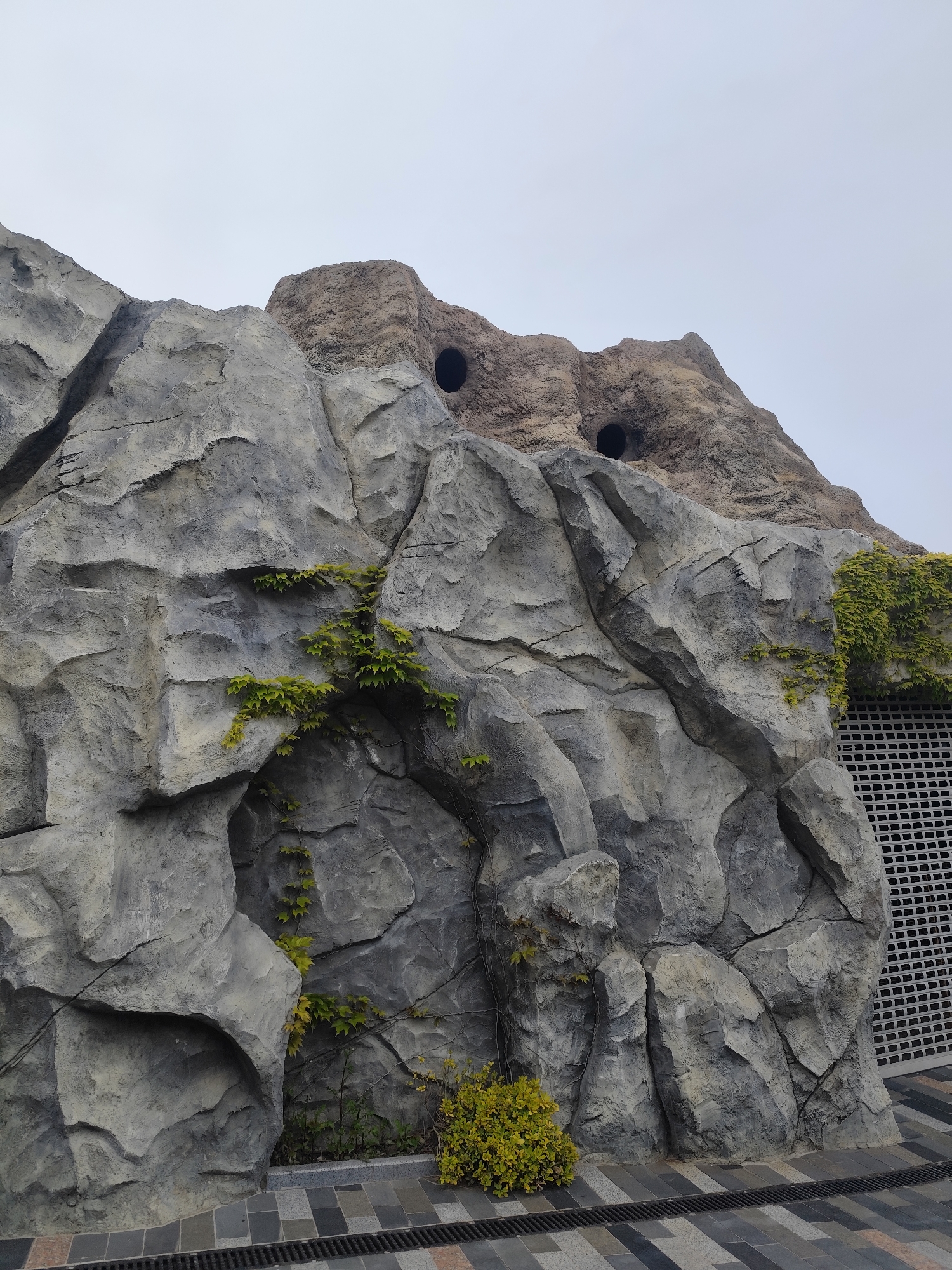 The image shows a faux rock formation next to a building in Симеизский поселковый совет, Ukraine. The rocks are gray and textured, with some greenery growing on them. The building behind the rocks has a gray grid pattern. The ground is tiled. The sky is overcast. There are two dark round holes in the top of the rock formation.