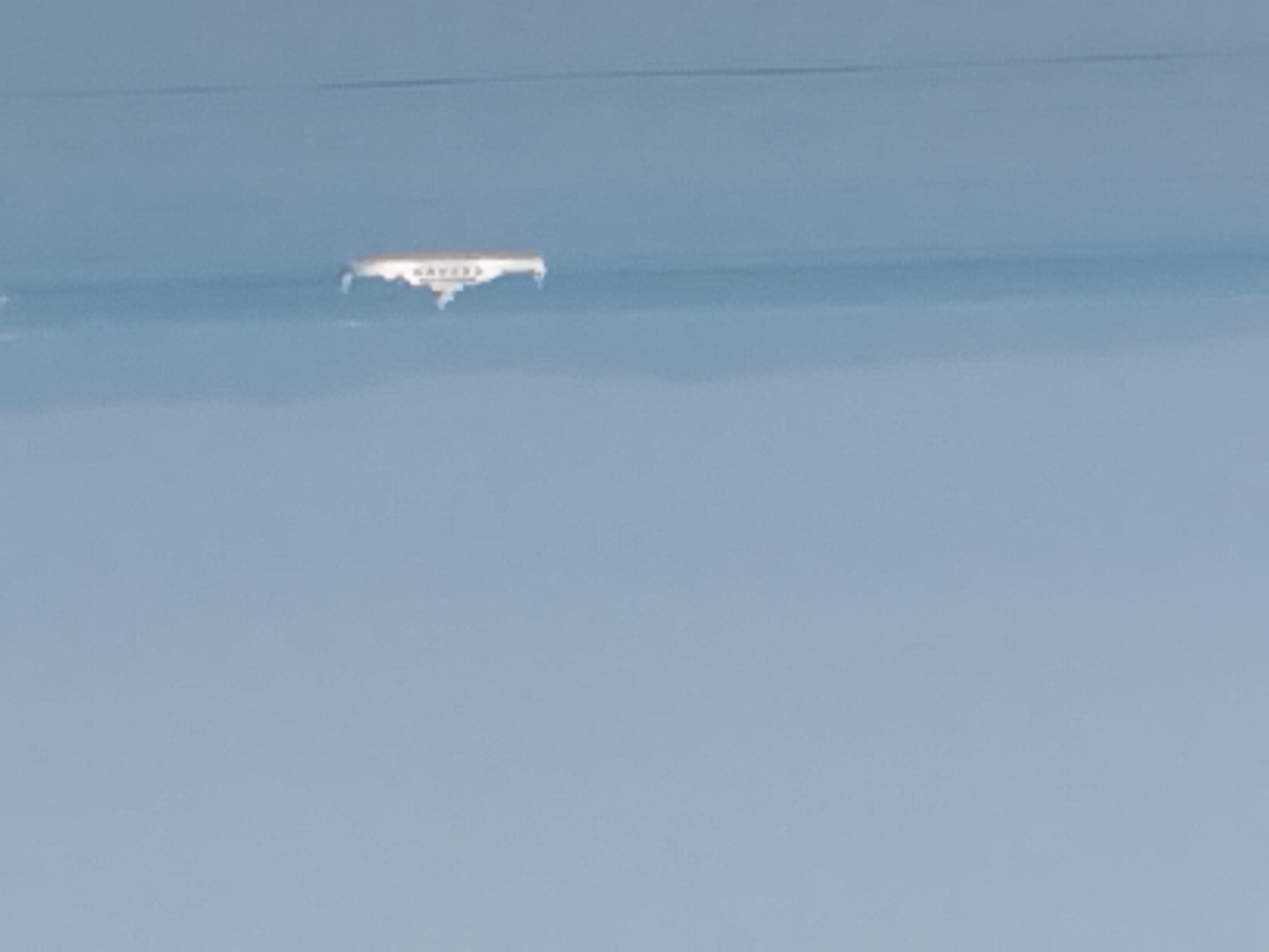 A ferry boat is visible in the distance on a calm body of water, likely the Adriatic Sea, with hazy landmasses in the background. The sky is a pale blue and appears overcast. The ferry is white with a yellow stripe along its side and a superstructure on top. The overall image is characterized by its soft focus and atmospheric perspective, suggesting a long-distance view. The scene evokes a sense of tranquility and open space.