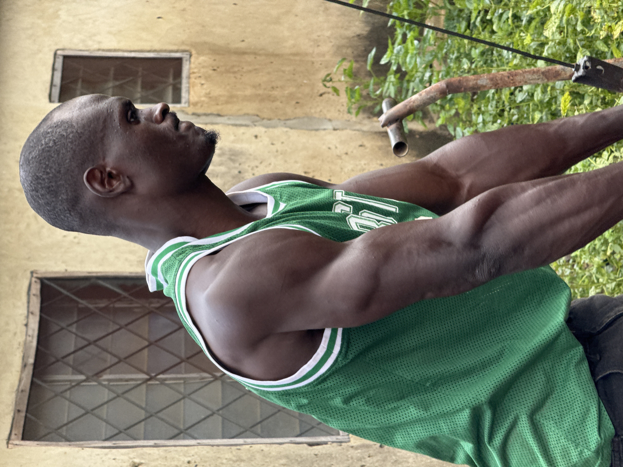 A medium shot captures a dark-skinned man from the chest up, wearing a green basketball jersey with white trim. He is looking to his right, with a slight furrow in his brow. The man has a shaved head and is sweating lightly on his forehead. His left arm and shoulder are prominent, showing well-defined muscles. Behind him, a beige wall with two windows is visible. The left window is larger and has a diamond-shaped grid pattern. The smaller right window is partially obscured. Green foliage and a rusty metal object are present in the background. The scene appears to be outdoors, possibly in a yard or alleyway, in the city of Akwanga, Nigeria.