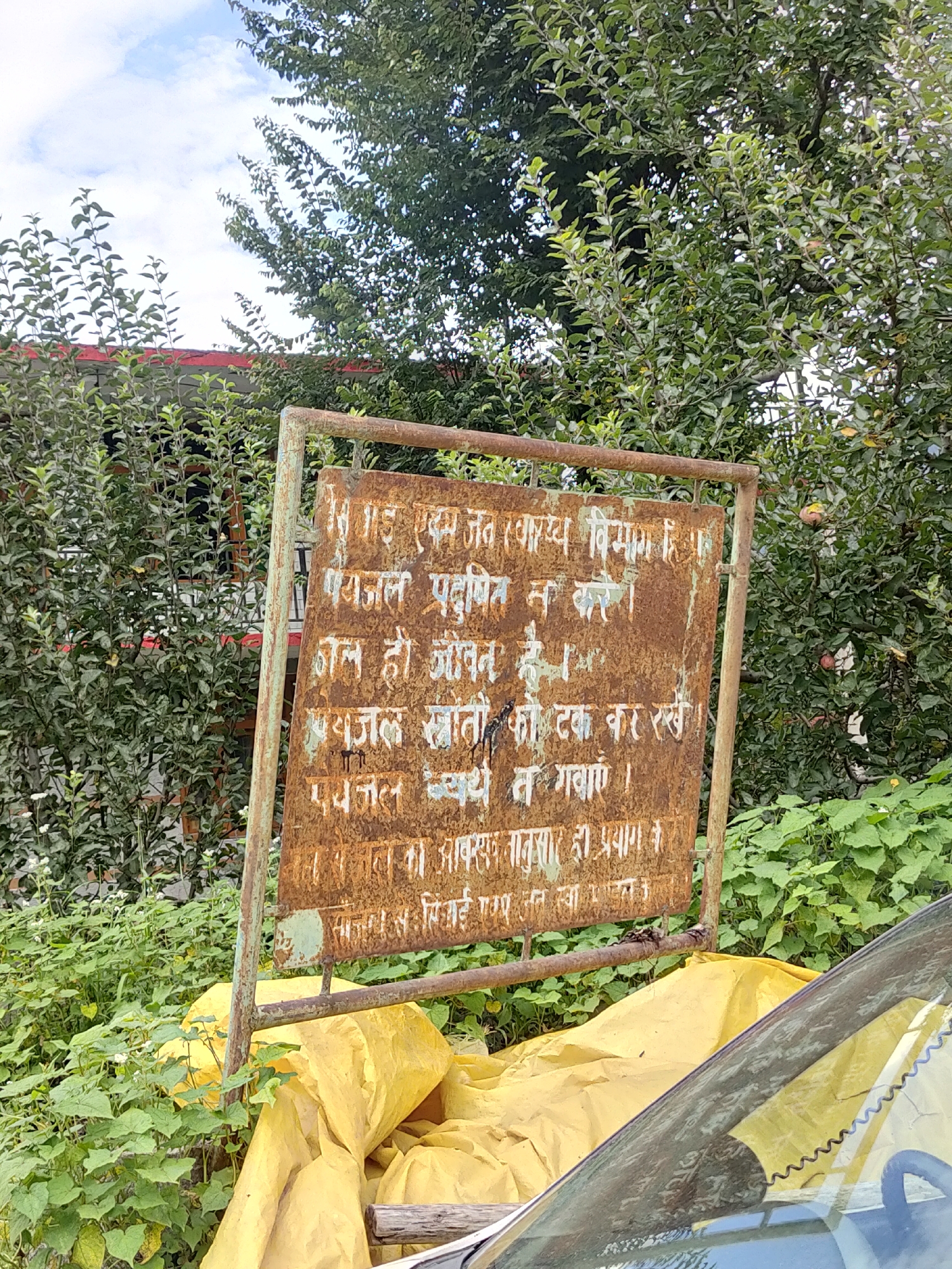 In Burwa, India, a rusted metal sign stands amidst lush green foliage. The sign, weathered and aged, displays text in Hindi. The first line appears to read "जित स्वास्थ्य विभाग हि"। The second line states "पयाजल प्रदूषित न करे"। The third line translates to "जल ही जीवन है"। The fourth line reads "पयाजल स्त्रोतो को टक्क कर रखे"। The fifth line conveys "पयाजल व्यर्थ न गबाएं"। The sixth line states "तदनानुसार ही प्रयाग"। The seventh line is "प्रयजन साई"। The sign is mounted on a rusty metal frame. In the foreground, a portion of a vehicle's windshield and a large yellow tarp are visible. The background is dominated by dense greenery, suggesting an outdoor, possibly rural or semi-urban setting. The sky above is partly cloudy. There are no people or animals clearly visible in the image.