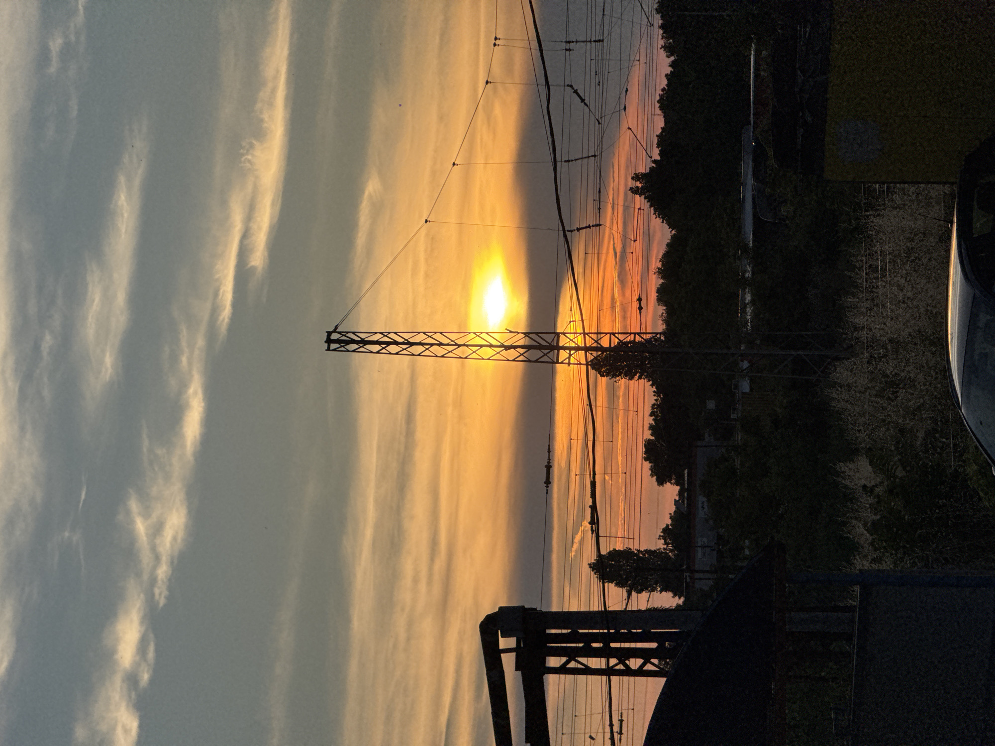 The image depicts a sunset over an industrial or railway area in Burgas, Bulgaria. The sky is filled with vibrant hues of orange, yellow, and gray from the setting sun, with wispy clouds stretching across. In the foreground, silhouetted against the colorful sky, are industrial structures, including a tall lattice tower supporting overhead power lines. Trees and vegetation form a dark line across the middle ground, with some of the foliage illuminated by the sunset. A dark, partially visible structure, possibly a building or a canopy, is on the left side of the frame, and a car is visible in the bottom right corner. The overall scene captures the dramatic beauty of a sunset in an urban industrial setting.