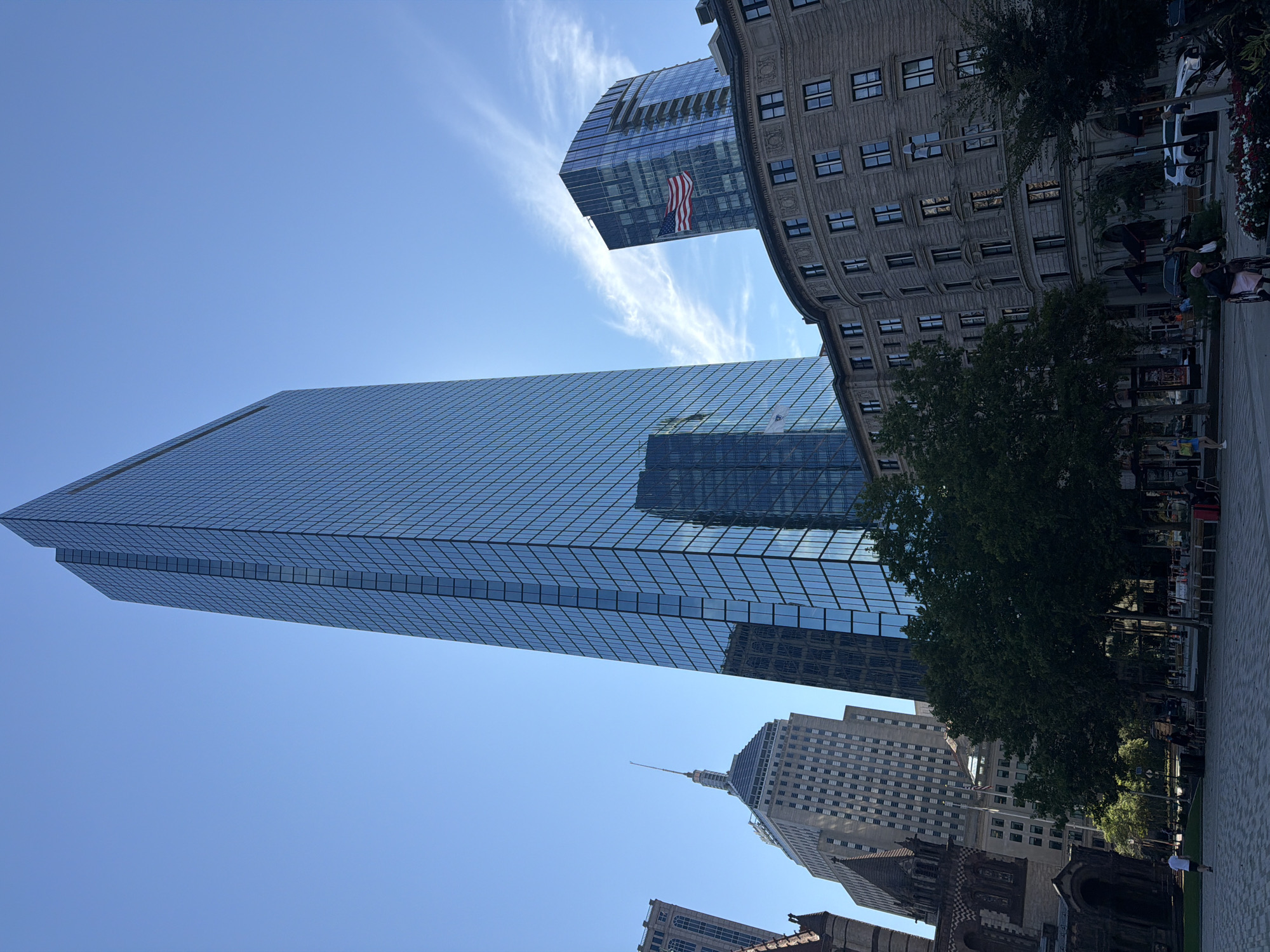 In Boston, Massachusetts, United States of America, a view of the city skyline is presented on a bright, clear day with a few wispy clouds. The John Hancock Tower, a prominent skyscraper, dominates the center of the frame with its sleek, glass facade reflecting the blue sky. To its right, another modern glass building displays the American flag. In the foreground, a curved, ornate stone building with many windows is visible, suggesting a historic district. Trees line the area in front of this building, and a paved plaza with benches occupies the lower part of the image. Several people are present, including a person in a wheelchair and others walking or sitting. Cars are parked along the street. The overall scene captures a mix of contemporary and historic architecture in an urban setting.