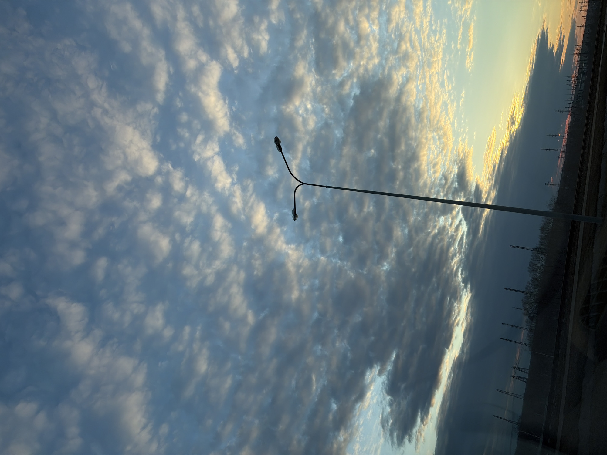 The image shows a sky filled with altocumulus clouds illuminated by the setting sun, casting a mix of blue and golden hues. In the foreground, a Y-shaped street lamp extends towards the right side of the frame. On the right edge of the image, a row of power line towers is visible against a darker sky and some vegetation. The location is identifiable as Chaikovsky, Russia by the characteristic power line towers and cloud formations. No people are visible. The scene appears to be taken from a low angle, possibly inside a vehicle.