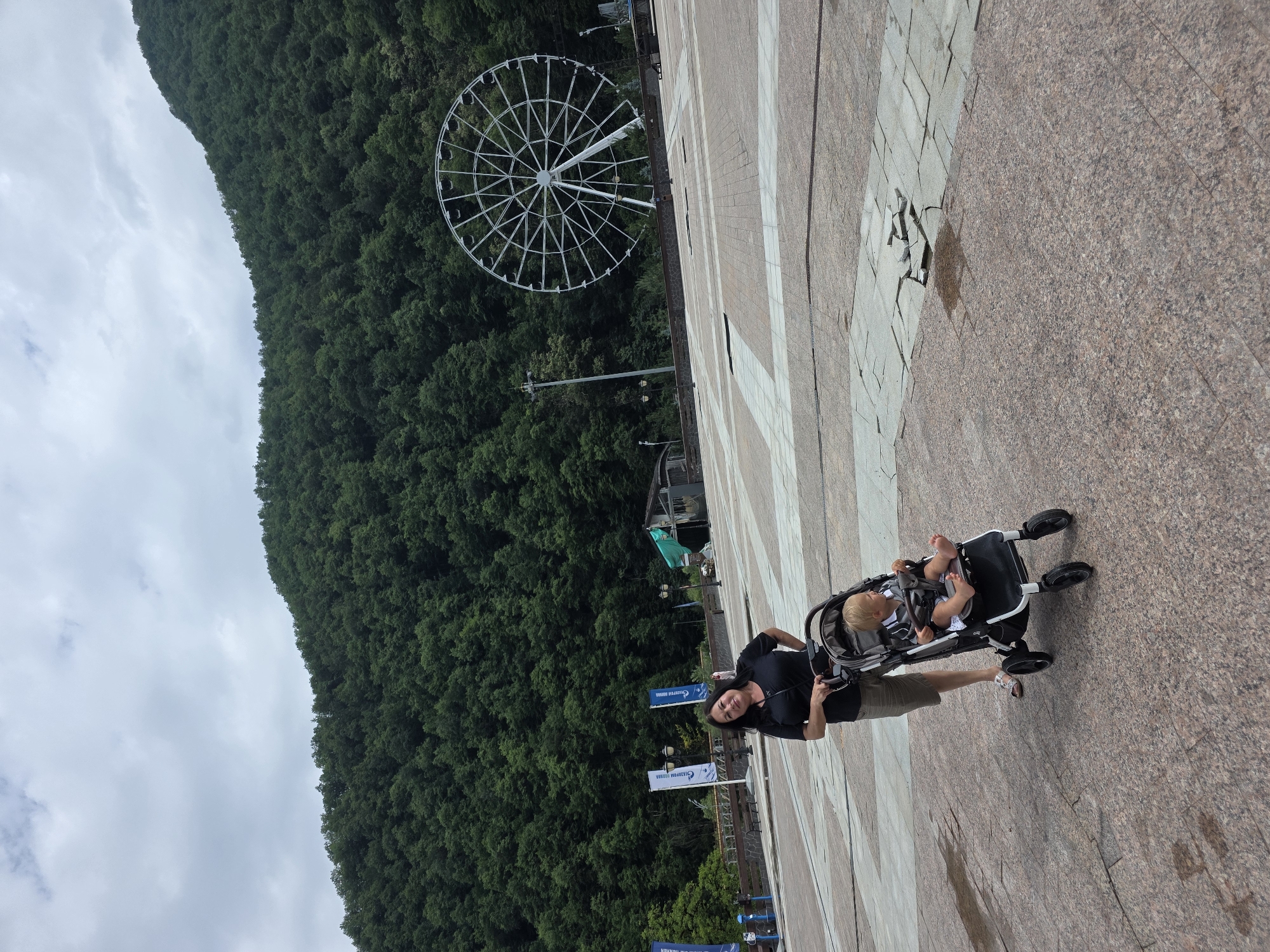 In Estosadok, Russia, a woman stands to the left of a stroller occupied by a young child. The woman is wearing a black t-shirt and shorts, with her left hand on the stroller handle. The child, with blonde hair, is looking forward, wearing a patterned shirt. Behind them, a large Ferris wheel is visible against a backdrop of a dense, tree-covered mountain under a cloudy sky. The ground is paved with large, square tiles, with some areas showing wear and damage. Flags and lampposts are also present in the background.