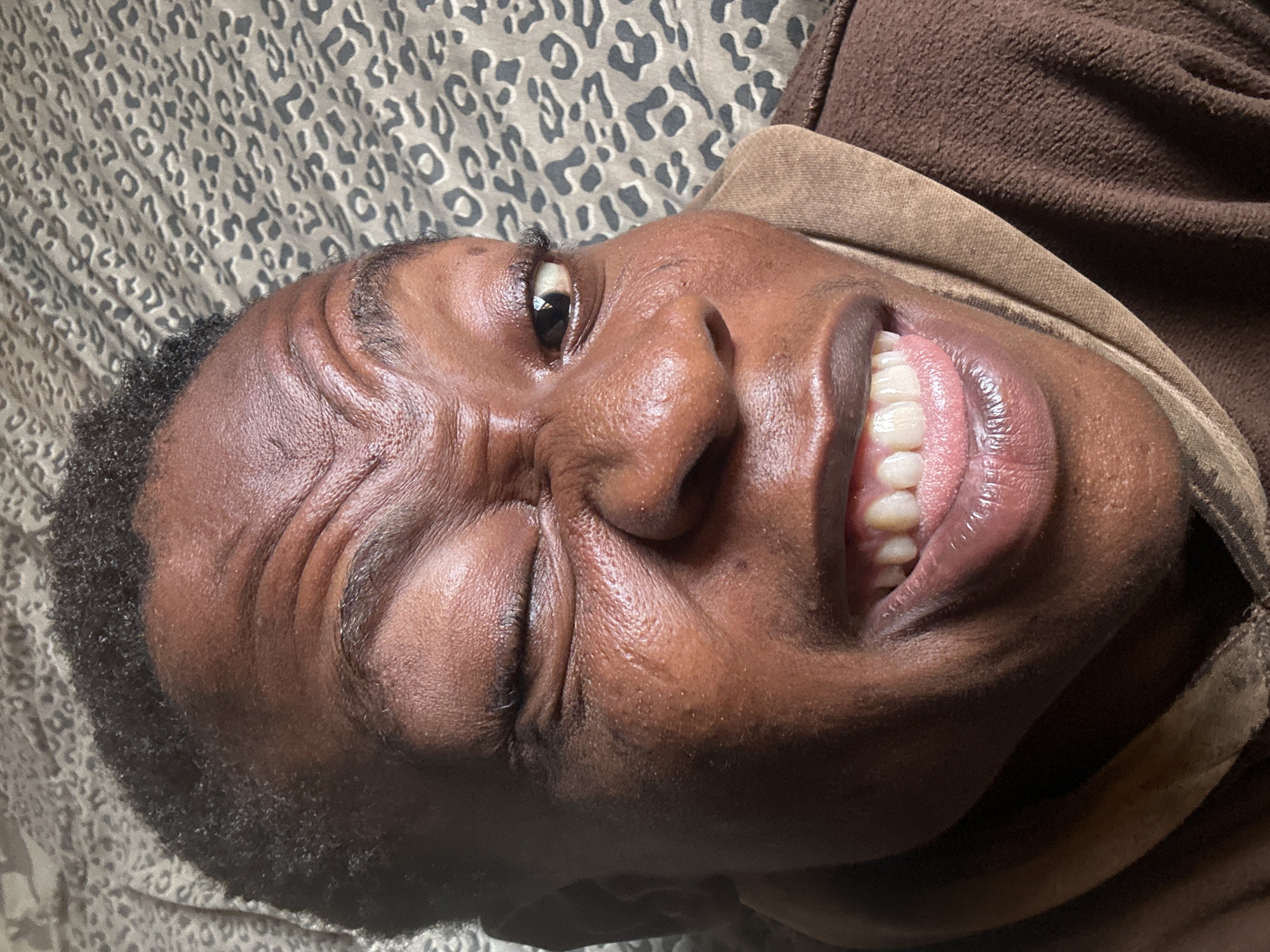 A medium close-up shot depicts a person with dark skin and short, curly black hair. The individual is lying down on a surface with a leopard print design. The person's face is contorted with the right eye squinted shut and the left eye partially open. The person is wearing a brown hooded sweatshirt. The location is unknown.