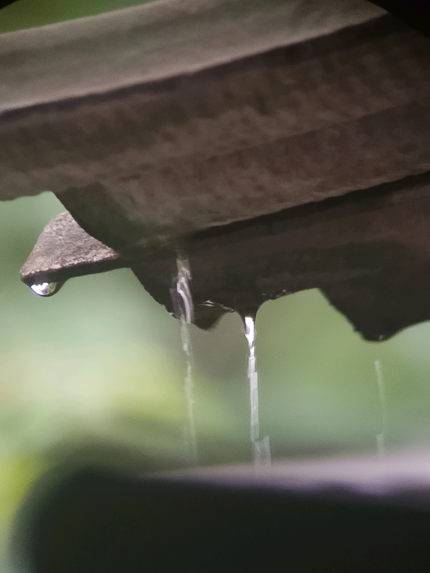 A close-up, macro shot captures water droplets falling from a dark, textured surface, possibly a roof or overhang. The background is a blurred, soft green, indicating an outdoor, possibly natural, setting. One prominent droplet is suspended mid-air, reflecting light, while others streak downwards as thin trails of water. The lighting is muted, suggesting overcast conditions or a shaded area. The scene evokes a sense of calm and the quiet beauty of nature. The image does not contain any specific landmarks or elements that would allow for the precise identification of a location within Jember, Indonesia.