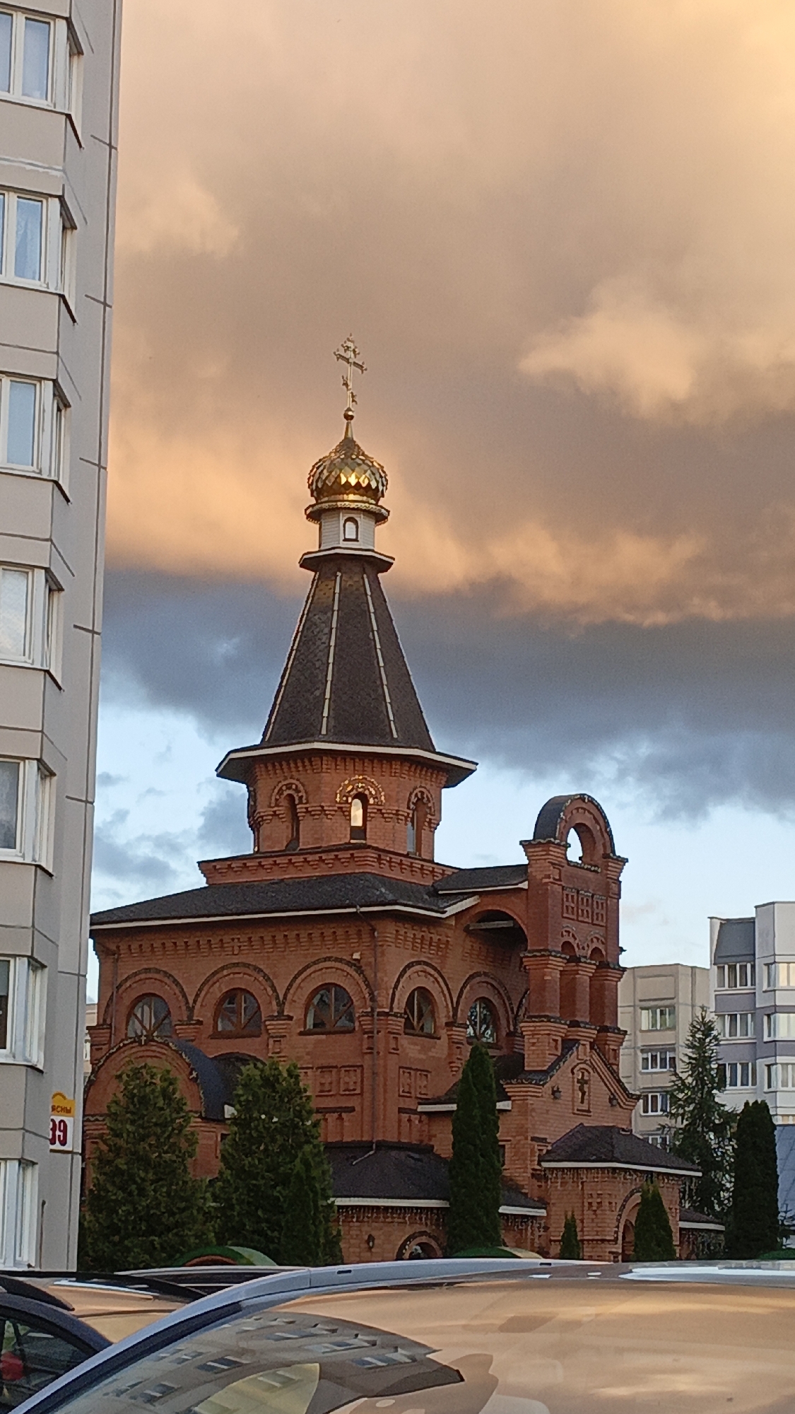 An exterior, medium-shot, vertical-orientation, eye-level, low-angle photograph captures an orthodox church constructed from red brick, seen from across a street with parked cars. The church features a golden dome topped with a cross, and a dark-colored, conical roof. The building has arched windows, decorative brickwork, and is flanked by trees. To the left of the church, a tall, light-colored apartment building with multiple windows is visible. In the background, other buildings and the sky with dramatic, overcast clouds illuminated by warm, sunset light are present. The scene appears to be set in the Бараўлянскі сельскі Савет, Belarus. There are no people or animals visible in the image.