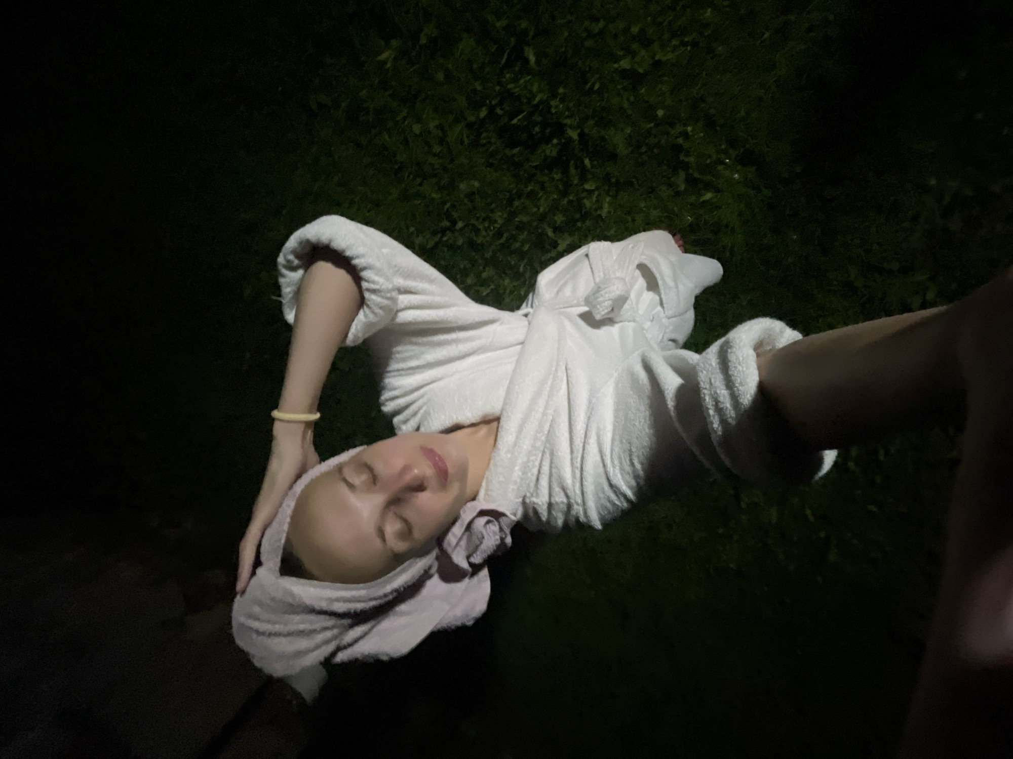 A woman, with her eyes closed and a serene expression, stands outdoors at night. She is wearing a white bathrobe and has a towel wrapped around her head. Her right hand is raised to her head, and her left arm is bent, holding the camera for a selfie. She appears to be in a grassy area, with dark shadows and what looks like foliage in the background. The lighting suggests it is nighttime, possibly illuminated by a single light source creating a spotlight effect on her. There is no visible information about the city of Sharkan, Russia.