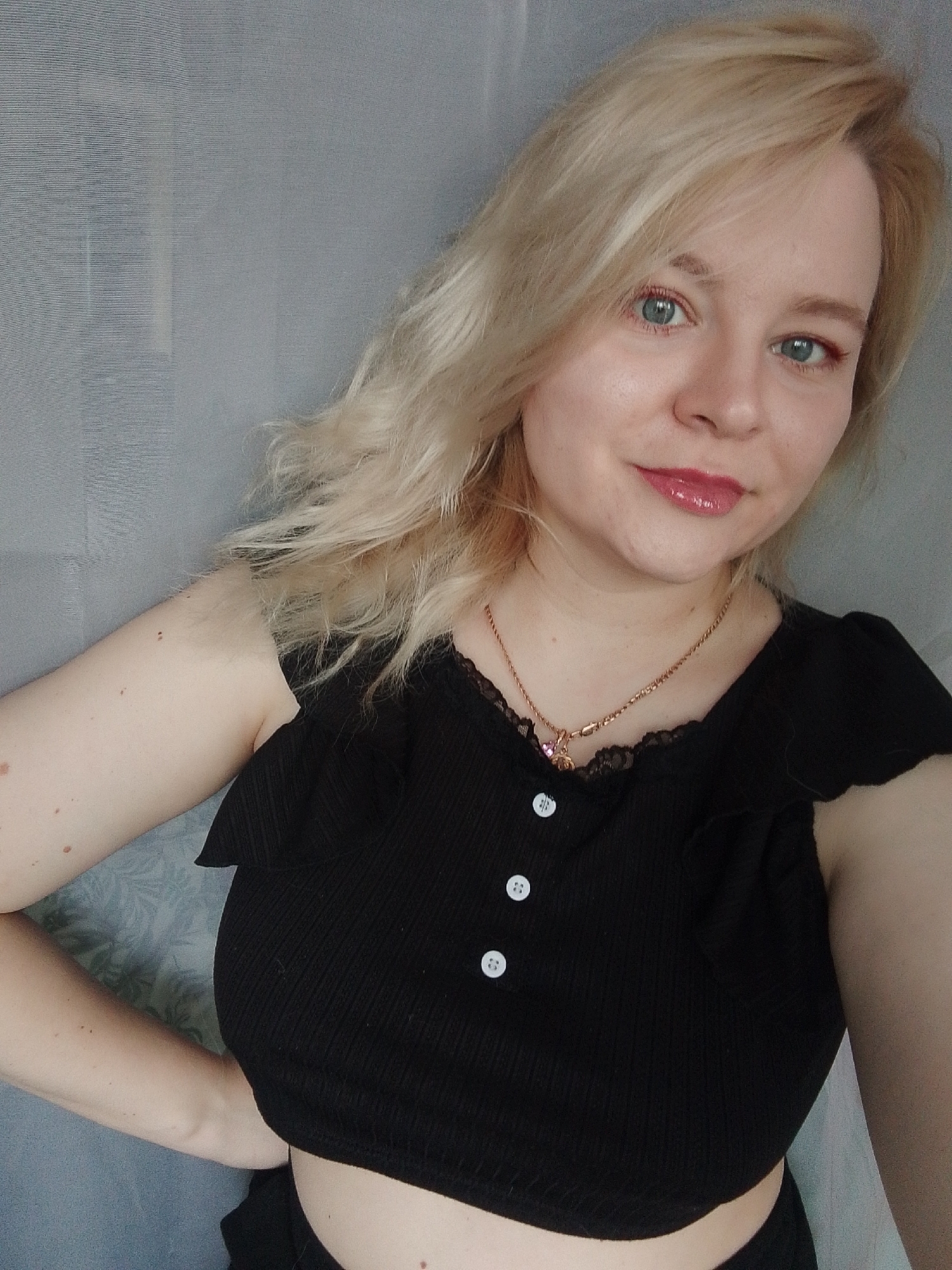 A woman with blonde hair is posing for a photo indoors. She is wearing a black cropped top with white buttons and a gold necklace. She has light skin and blue eyes. She has light makeup on. The background is a light gray wall with a subtle floral pattern. The location cannot be determined to be in Izhevsk, Russia from the image.