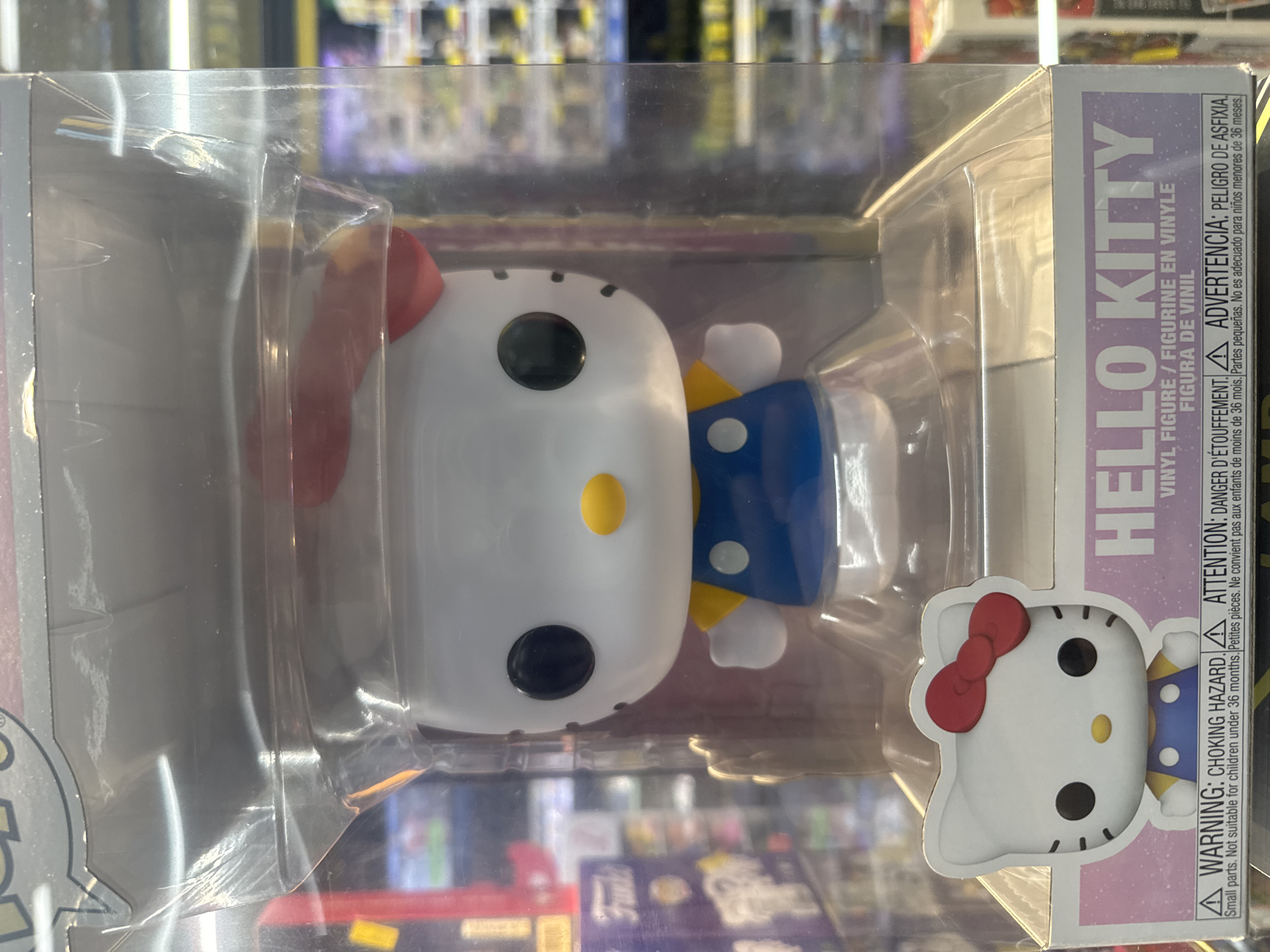 A Funko Pop! vinyl figure of Hello Kitty is displayed in its original plastic packaging. The figure is white with black eyes, a yellow nose, and a red bow on its head. It wears a blue dress with a yellow collar and white buttons. The packaging is clear plastic with some scuff marks and creasing visible on the upper portion. A warning label in multiple languages is affixed to the bottom of the packaging. The background is blurred, showing shelves with other merchandise. The image appears to be taken in a retail store environment. The specific location in Budva, Montenegro, cannot be determined from the visual information provided.