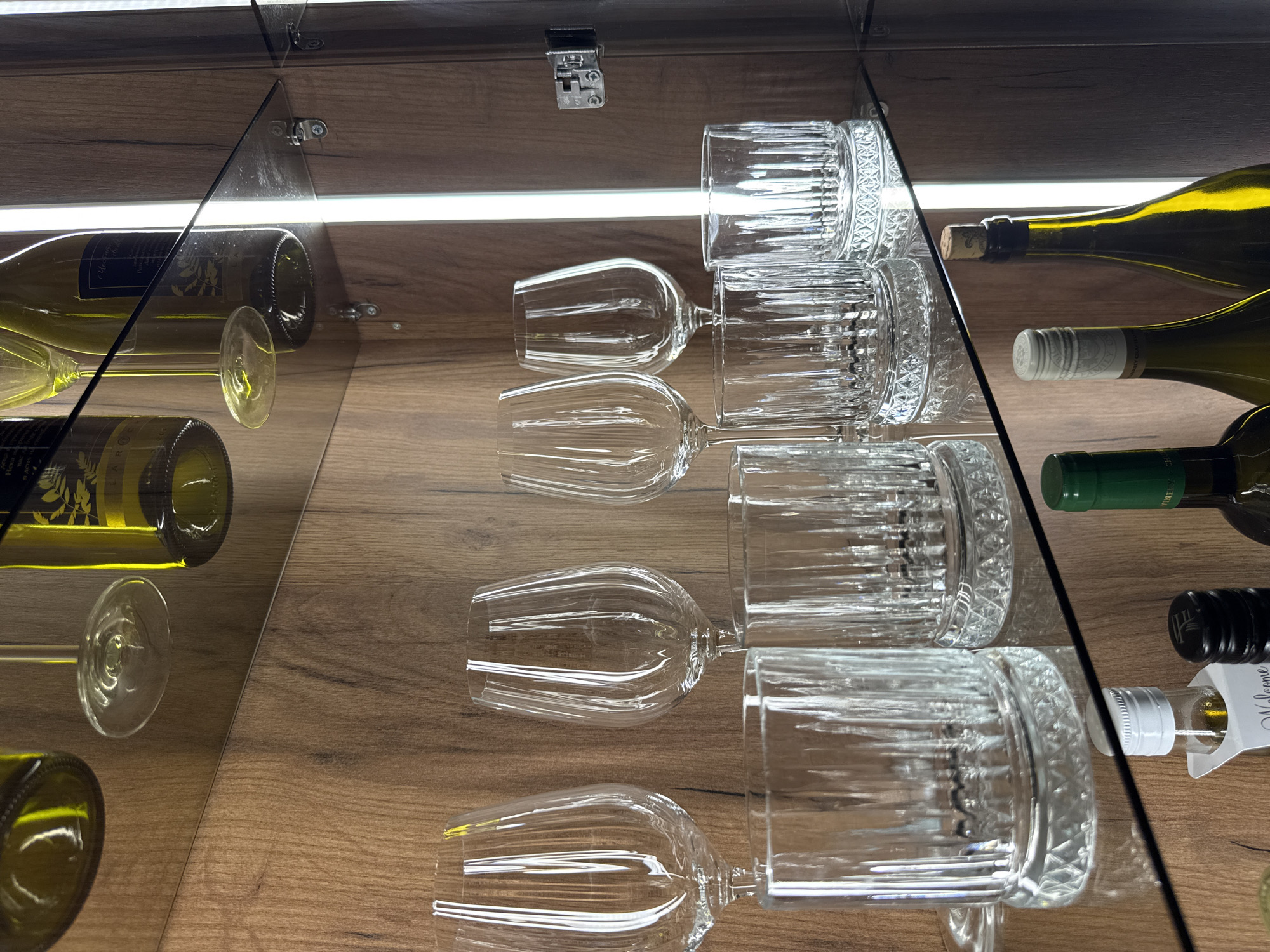 The image shows a well-lit display cabinet with various glassware and bottles. On the top shelf, several wine bottles, some partially visible, are placed upside down, suggesting they are part of a decorative display or storage. Below this, on a glass shelf, are several wine glasses and short tumblers, also clear glass and appearing to be clean and unused. Further down, on another glass shelf, are more tumblers and two wine bottles, one with a cork closure and the other with a screw top. A small vial with a white cap and some liquid is also visible. The cabinet interior has a wood-grain finish and is illuminated by a vertical light source on the right side. The scene appears to be an indoor setting, possibly a home bar or a retail display for beverages. There is no indication of the image's location in Plovdiv, Bulgaria.