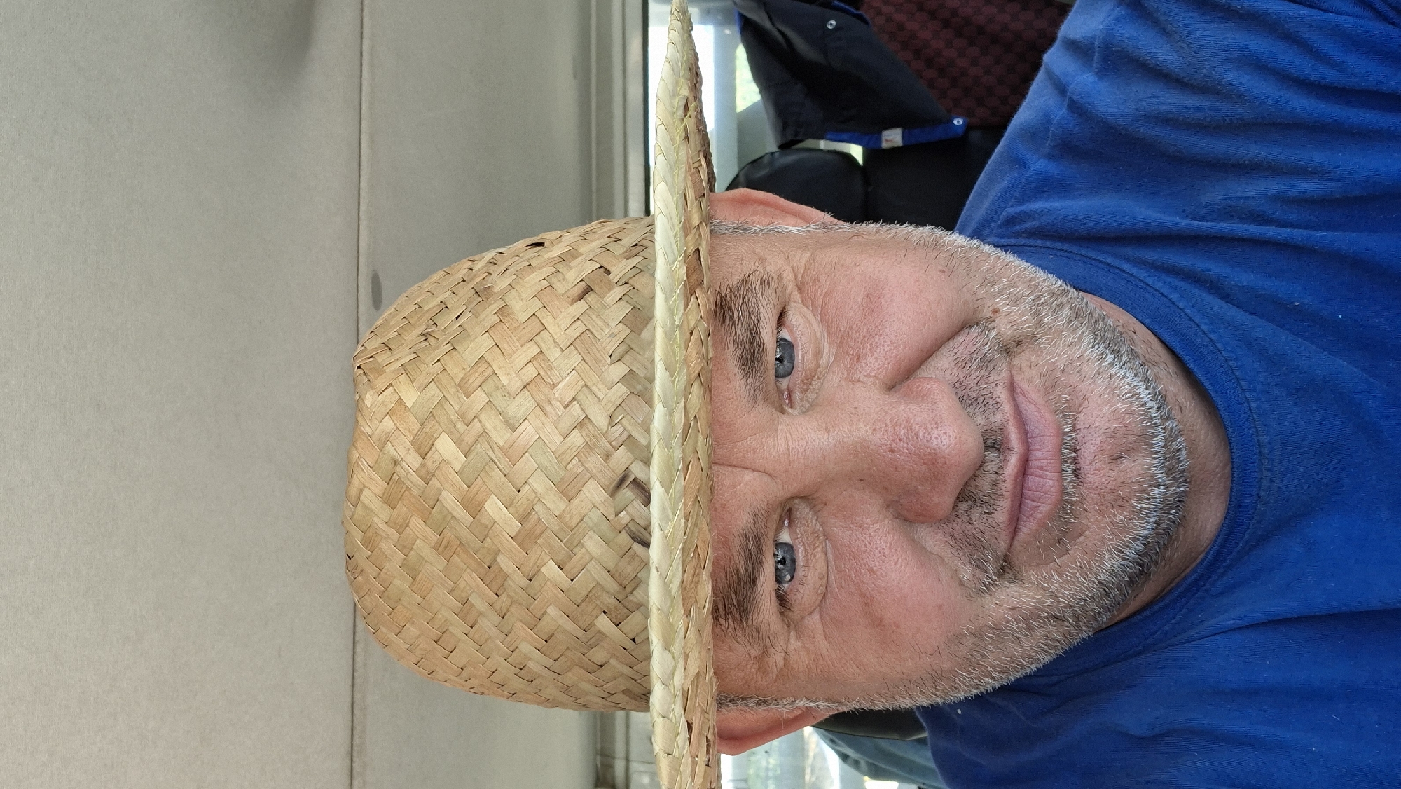 A close-up, head-and-shoulders selfie shows a man wearing a straw hat and a blue t-shirt. He is in the front seat of a vehicle, with the interior of the car visible in the background, including the headliner and seats. The man has blue eyes, fair skin, and a light stubble beard. The lighting suggests it is daytime, possibly in Hîncești, Moldova.
