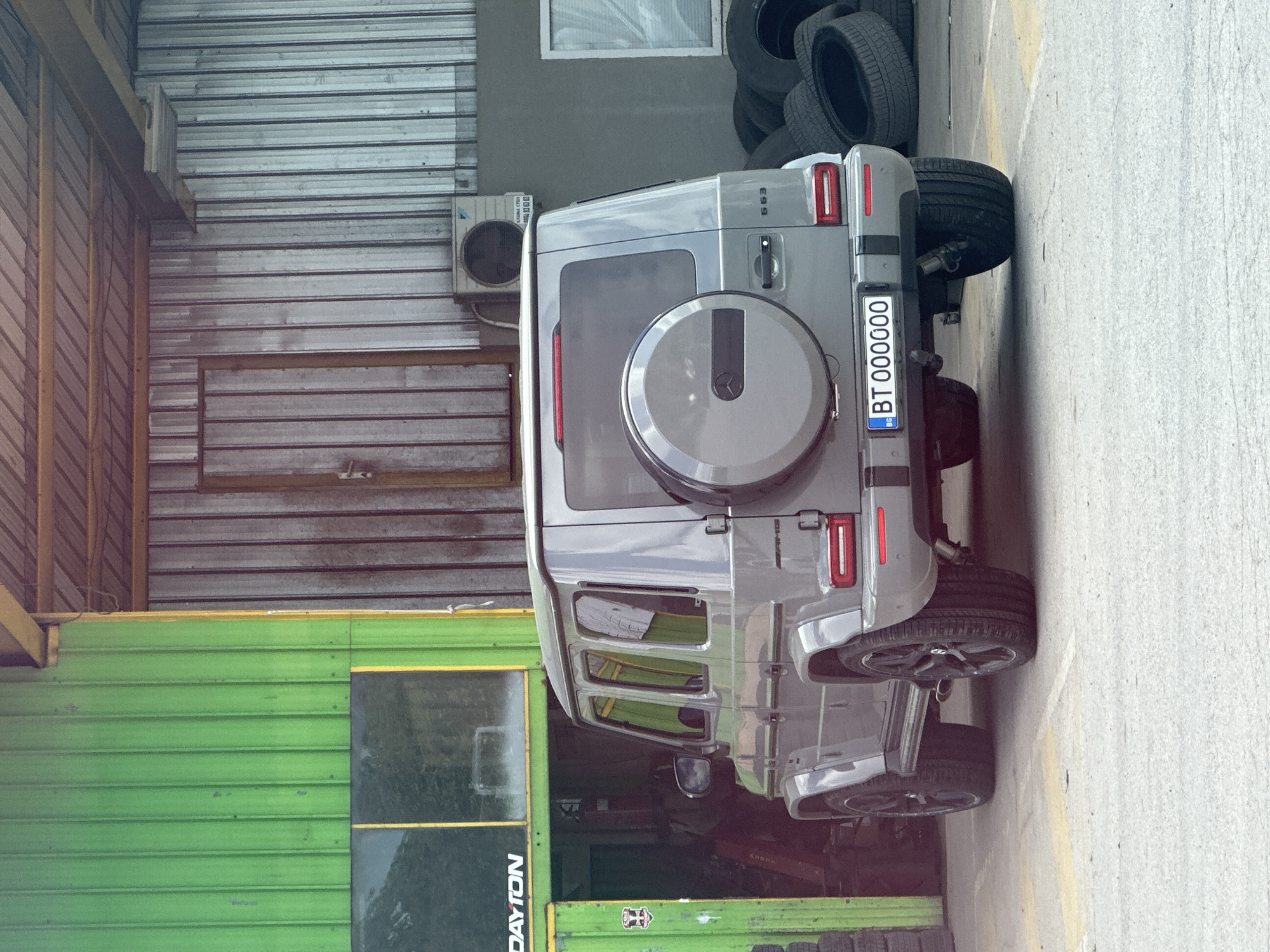 The image shows a gray Mercedes-Benz G63 AMG parked in an urban setting. The car's license plate reads "BT 000000," indicating it is registered in the Veliko Tarnovo region of Bulgaria. The vehicle is positioned on a concrete surface, possibly a parking area or a street. To the left of the car is a building with both green and gray siding and a "DAYTON" sign. Behind the car, against the back wall, are stacks of tires. An air conditioning unit is mounted on the gray portion of the wall. The scene appears to be near an automotive service or tire shop.