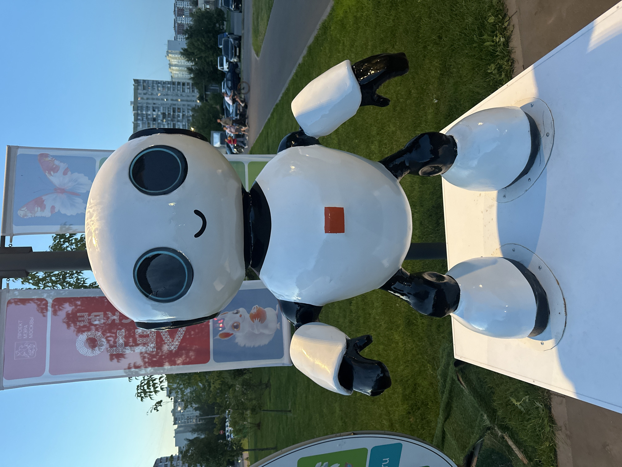 An outdoor, eye-level, medium shot captures a white and black robot sculpture, roughly two meters tall, standing on a white platform. The robot has a large, round head with two prominent black and teal eyes and a small, curved mouth. Its body is bulbous, with a small red square on its chest. The robot's arms and legs are black and white, with black hands and feet. Behind the robot, a lamppost supports two banners. The left banner is red and features the Moscow coat of arms and text in Russian that reads "ПРОЕКТ МЭРА МОСКВЫ" (Mayor's Project of Moscow). The right banner displays a picture of a butterfly. Further in the background, green grass, trees, and several multi-story apartment buildings are visible under a clear blue sky. A paved path runs alongside the grass, and cars and a person on a bicycle are present on the path, indicating a public urban space. The overall scene suggests an art installation or public display in a residential area of Moscow.