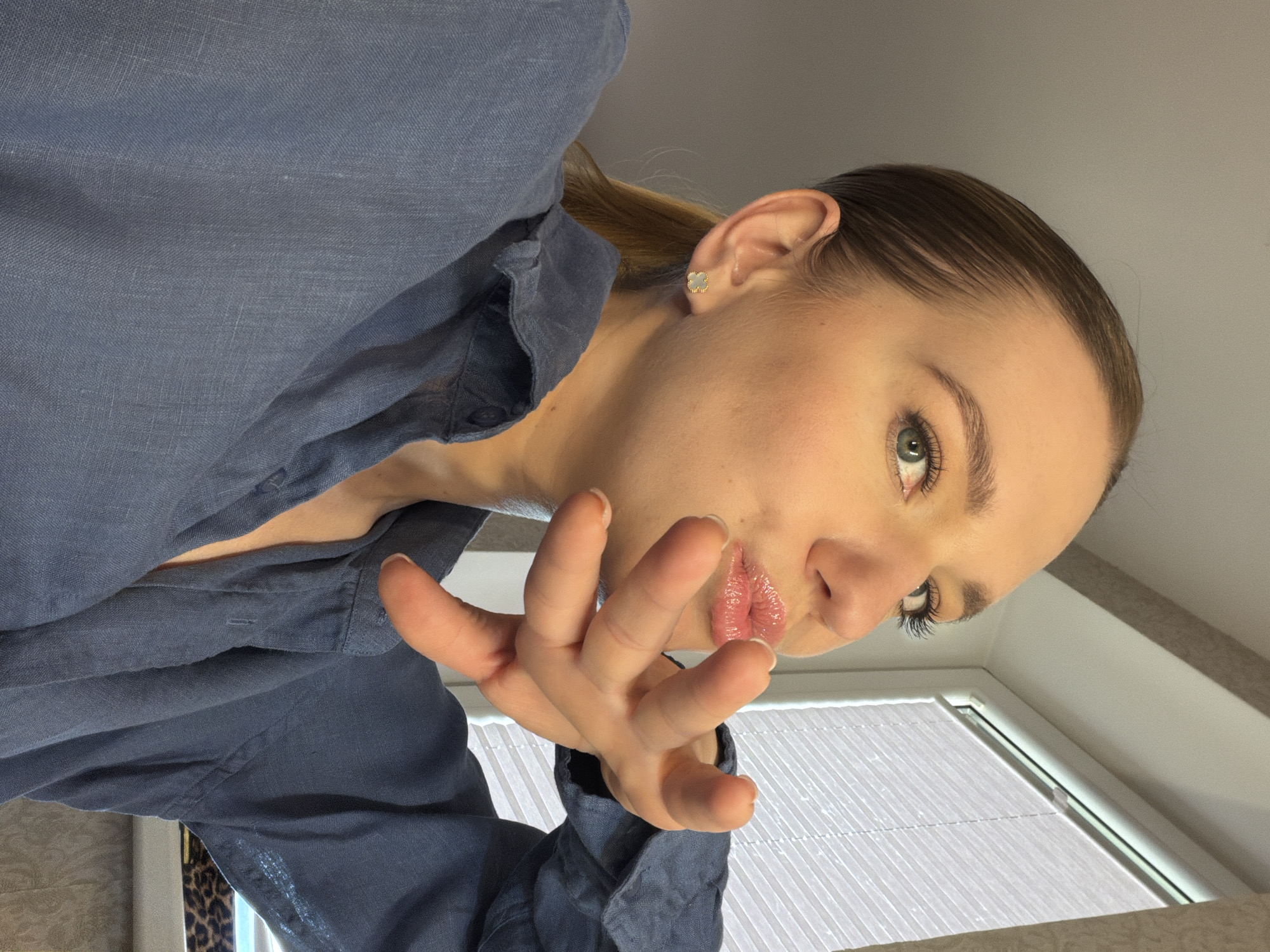 A low-angle, indoor portrait captures a young woman from Dreiliņi, Latvia, looking directly at the camera. Her light brown hair is pulled back, and she has fair skin, prominent eyelashes, and lips adorned with gloss. A small, light-colored, possibly clover-shaped earring with a gold outline is visible in her right ear. She is wearing a casual blue button-up shirt, appearing to be made of linen, with some buttons unfastened. Her right hand is raised towards the viewer, fingers slightly curled in a gesture that suggests engagement or invitation.
The background primarily features a white window with horizontal blinds, indicating an interior setting. Parts of a plain wall or ceiling are also visible. The lighting suggests daytime. The woman's expression is direct and appears confident or playful. There is no visible text in the image.