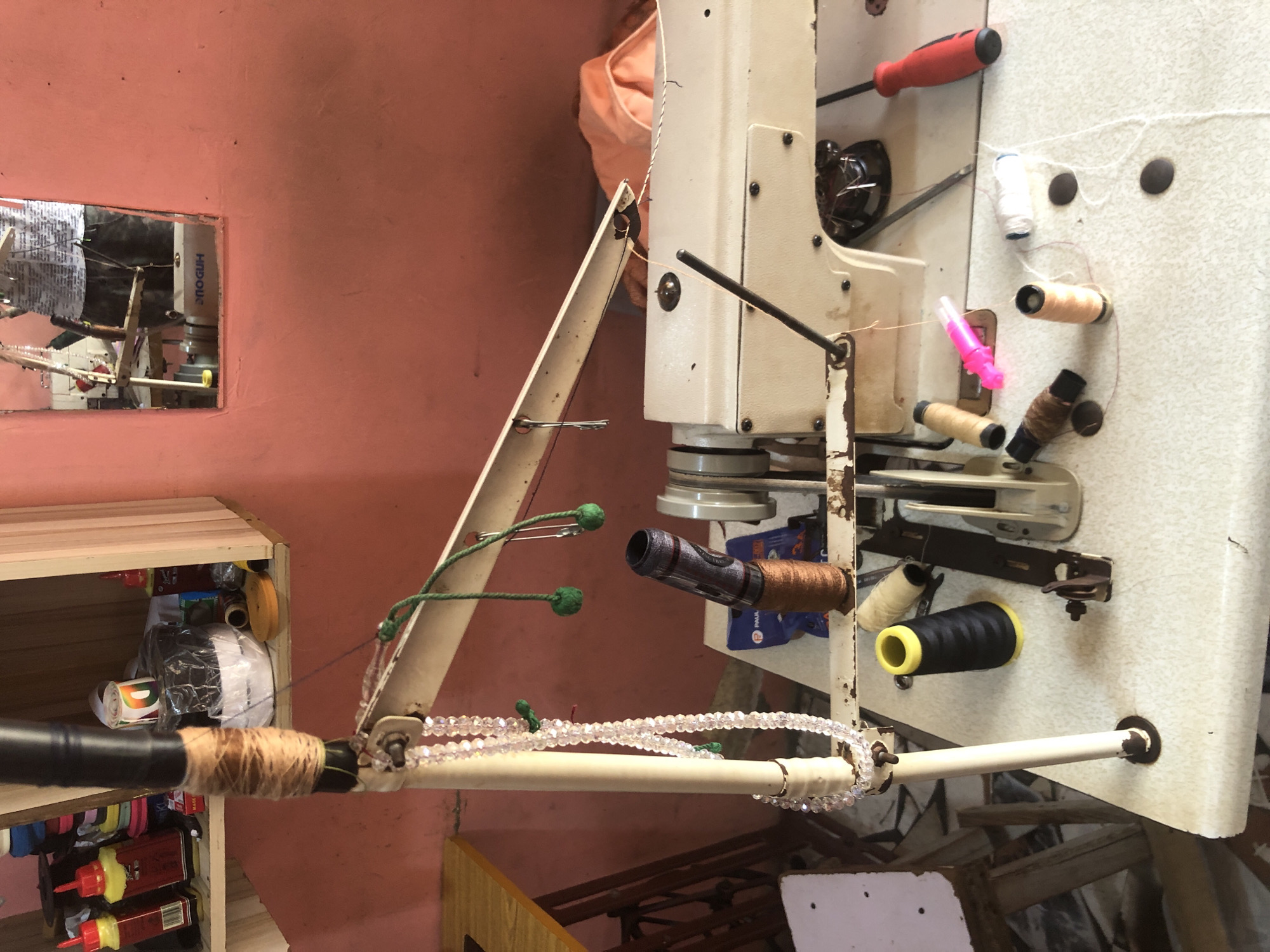 The image depicts a sewing machine in what appears to be a small workshop or home sewing area. The sewing machine is a vintage, cream-colored model with various spools of thread, bobbins, and sewing accessories scattered on the table around it. A beaded string hangs from a stand next to the machine, and a safety pin is attached to a green string, also hanging nearby. In the background, shelves are stocked with various items, including bottles of what seem to be adhesives or lubricants and rolls of fabric. A mirror on the wall reflects a person, partially obscured, who is likely operating or interacting with the sewing machine. The environment suggests a functional, working space, possibly in a less developed or more traditional setting. The overall impression is one of a small-scale tailoring or repair business.