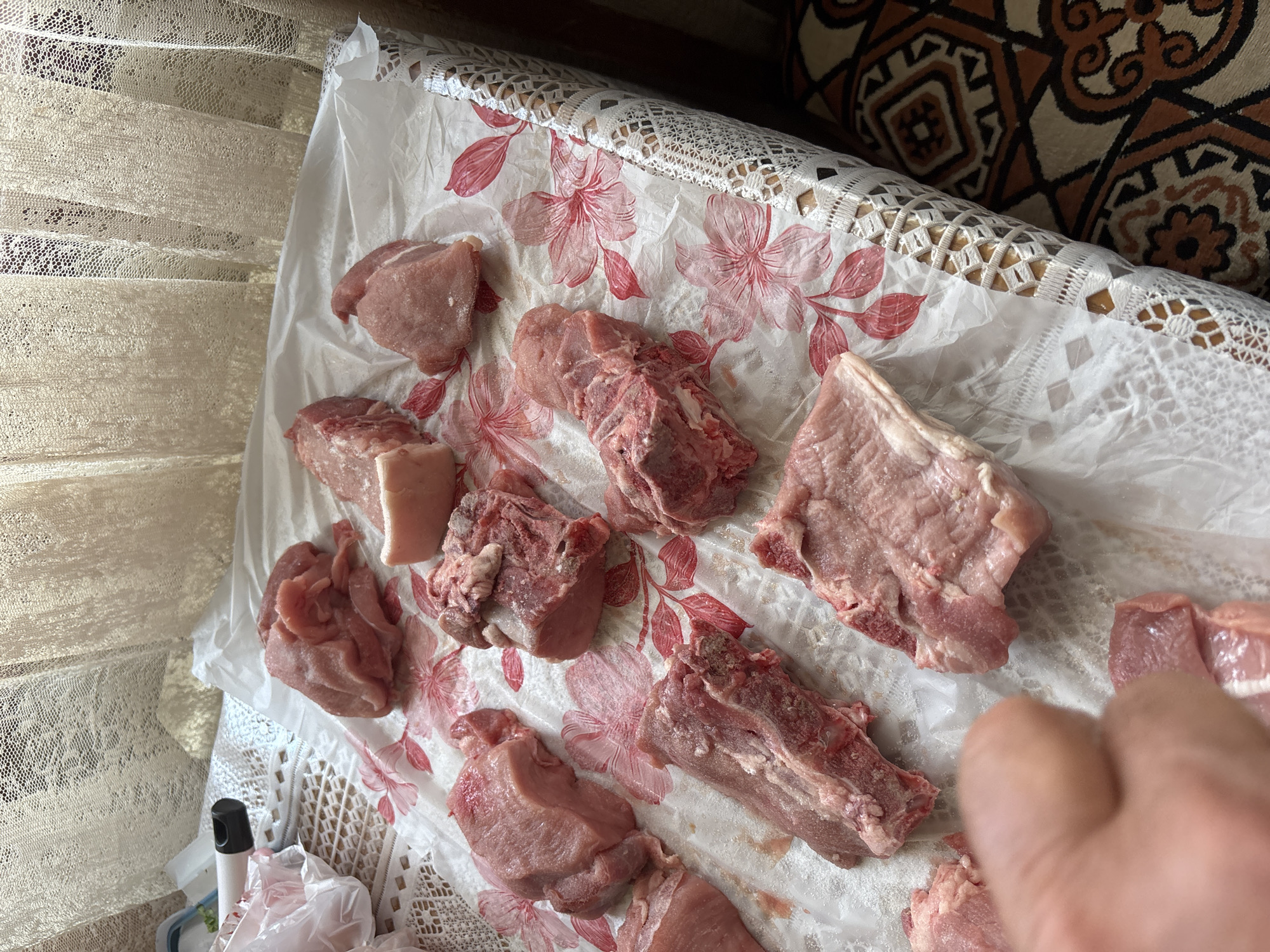 The image captures an indoor scene depicting the preparation of raw meat. Various cuts of pink raw meat, some with visible bone and fat, are laid out on a white plastic sheet. This sheet features a distinctive red floral pattern, primarily of large flowers and leaves. Beneath the patterned plastic sheet, a white, intricately woven or crocheted lace-like fabric is visible, likely serving as a tablecloth.
In the upper right background, a decorative cushion or textile with a brown, white, and beige geometric pattern is partially visible, suggesting an ethnic or traditional decor style, consistent with the given location context of Vagharshapat, Armenia. To the far left, a window provides natural light, and is partially covered by sheer, light-colored lace curtains.
A person's hand, with fingers curled, is visible in the bottom right corner, suggesting active involvement in handling or arranging the meat. In the lower left, a white spray bottle with a black cap and a crumpled clear plastic bag are present, hinting at food preparation or cleaning activities. The scene appears to be set during the daytime due to the bright, natural illumination. No discernible text is visible on any objects. The overall impression is one of domestic food preparation.