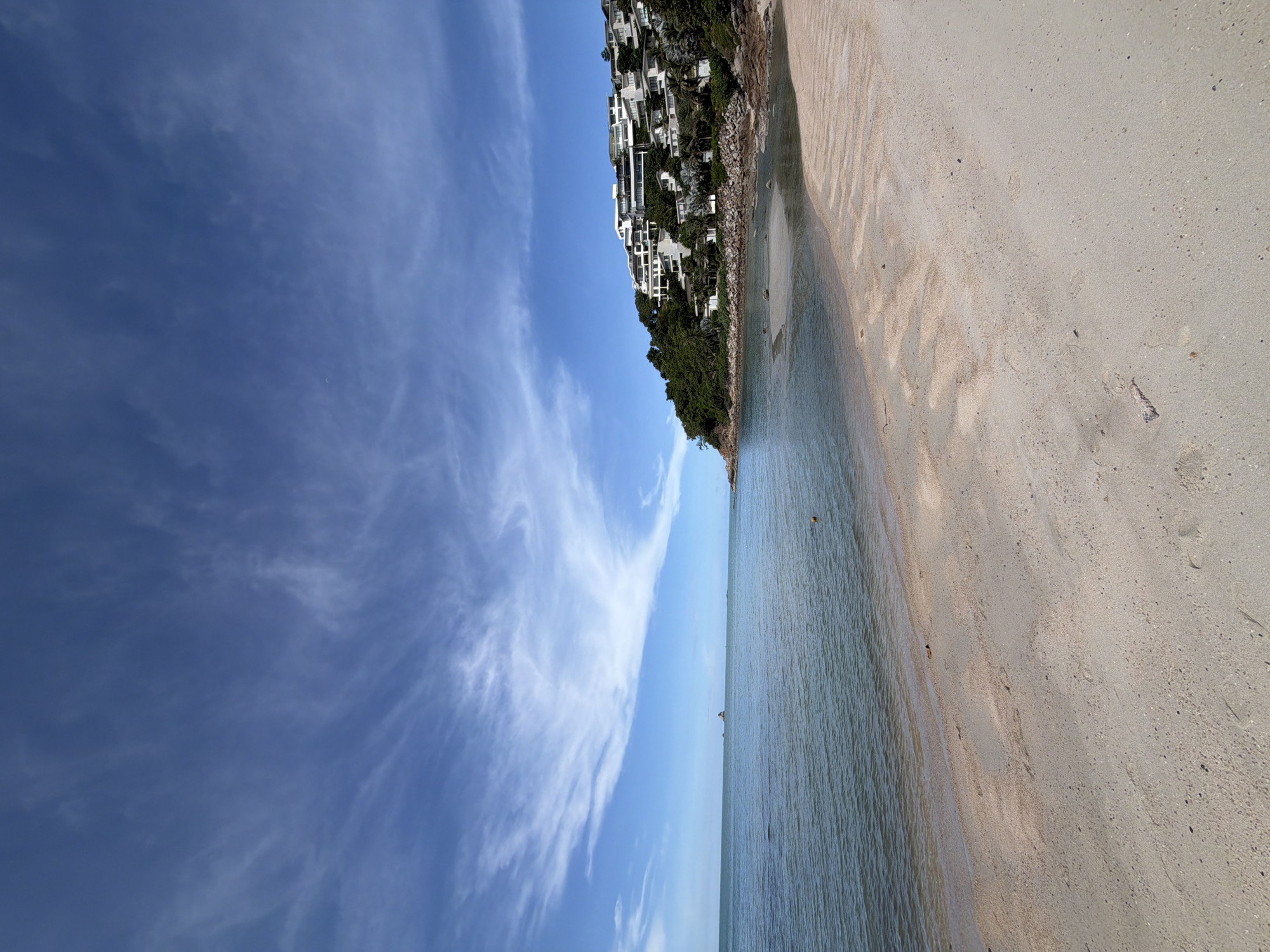 A wide-angle, eye-level shot captures a tranquil beach scene in Baan Plai Laem, Thailand. The foreground is dominated by a light-colored sandy beach, marked with subtle imprints and a few small pieces of debris. The sand transitions to a calm, turquoise ocean that gently laps at the shore. In the distance, a small, rocky islet stands in the sea.
Along the right side of the frame, a lush, green hillside rises steeply from the water. Dotted across this hillside are modern, multi-story white buildings, characteristic of a resort or residential complex. The architecture is clean and contemporary, with balconies and large windows facing the sea. Scattered among the buildings and trees are rocks and boulders.
The sky above is a vibrant blue, adorned with wispy white cirrus clouds that stretch across the expanse, creating a dynamic yet serene atmosphere. The overall impression is one of natural beauty and luxurious coastal living.