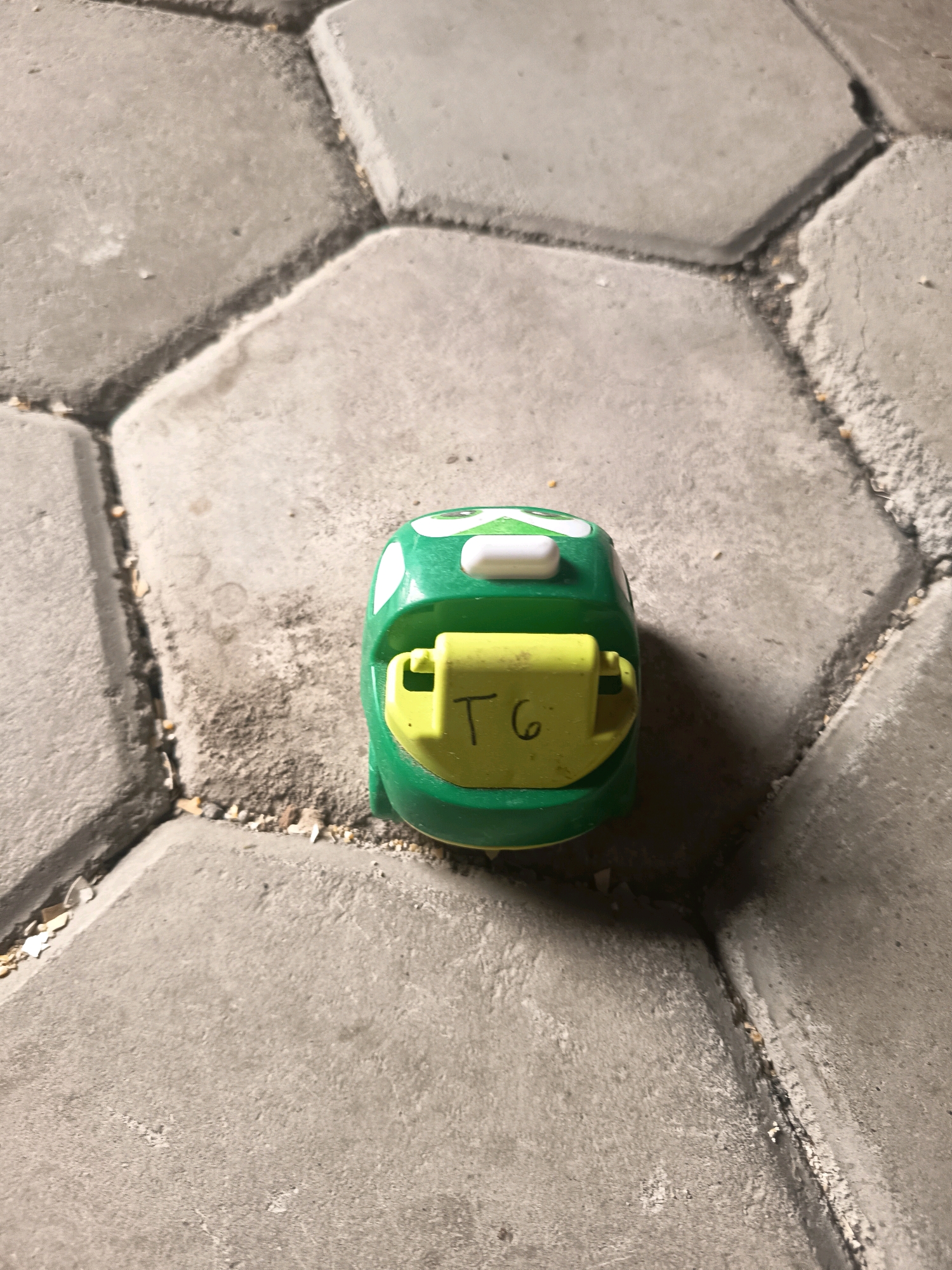 A green toy car with white accents on its headlights sits on a gray, hexagonal paved surface. A yellow rectangular piece with the letters "T6" written in black marker is attached to the front of the car. The paved surface is cracked and appears weathered. The background consists of more of the same paving stones. The scene is outdoors, likely in an urban environment.