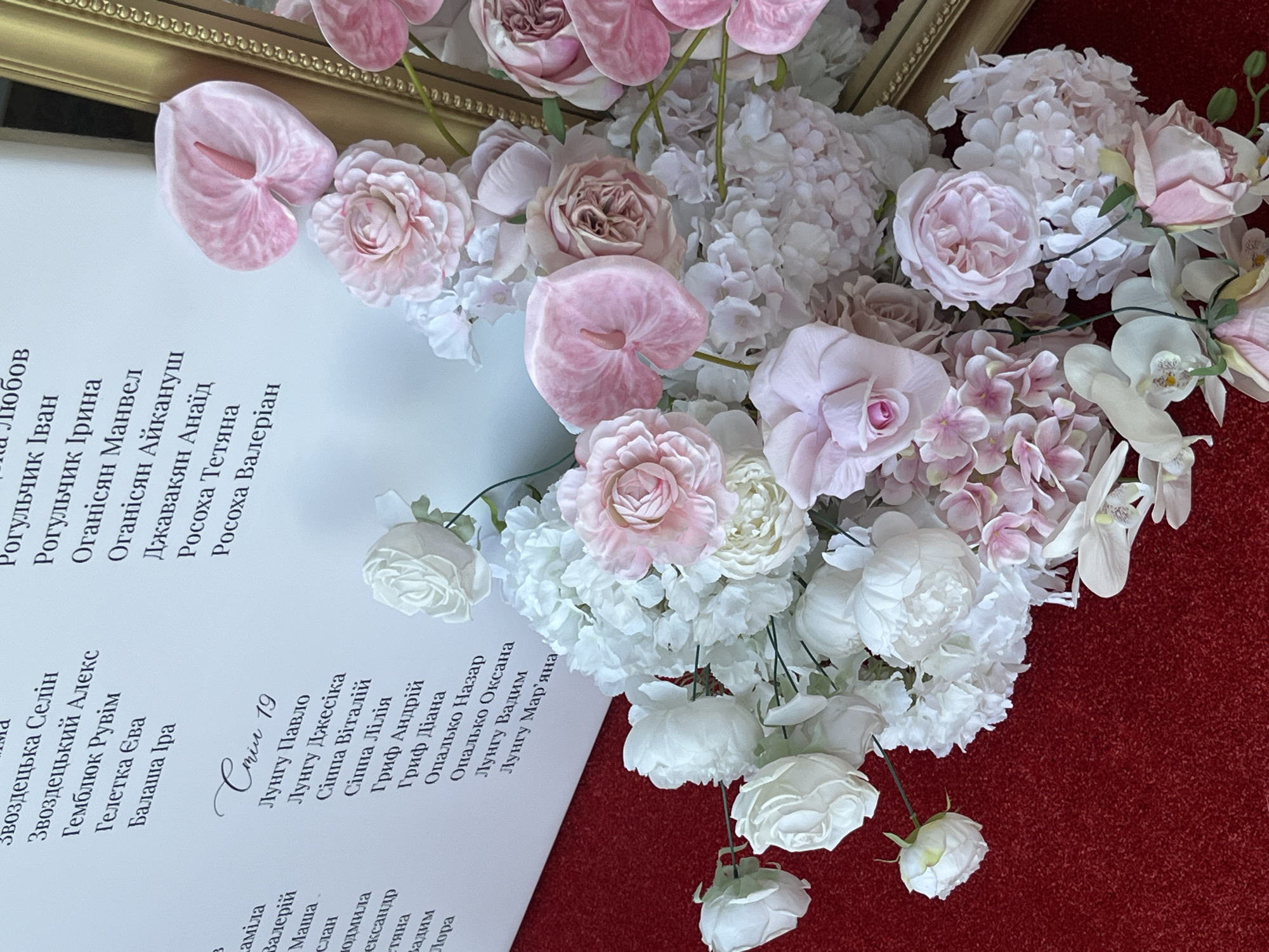 An indoor, eye-level, medium shot captures a close-up of a decorative floral arrangement and part of a white board displaying text in Ukrainian. The board is positioned on the right side of the frame, leaning slightly forward, and is covered in an elegant script. To the left of the board, the floral arrangement is densely packed with various artificial flowers in shades of pink and white, including roses, hydrangeas, and calla lilies. The arrangement rests on a textured red surface, likely a tablecloth or carpet. The overall scene suggests a formal event, potentially a wedding or banquet, in the village of Dubivtsi, Ukraine. The lighting is soft and diffused, creating a gentle illumination that highlights the textures of the flowers and the lettering on the board. No people are visible in the frame.
