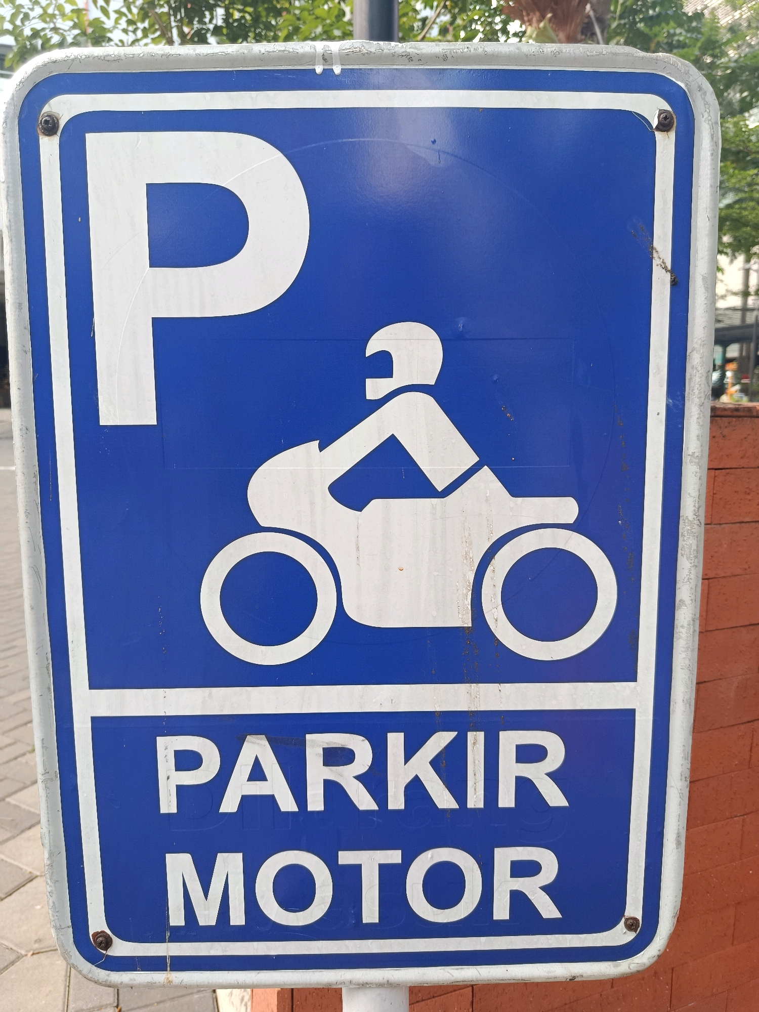 A blue and white rectangular sign is affixed to a pole. The sign has white text and a white graphic. The text at the top reads "P" in a large font. Below that is a graphic of a person riding a motorcycle. Below the graphic, in white text, are the words "PARKIR MOTOR". The sign is in a city setting. The background includes trees, a sidewalk, and a brick wall. The sign appears to be located in South Jakarta, Indonesia.