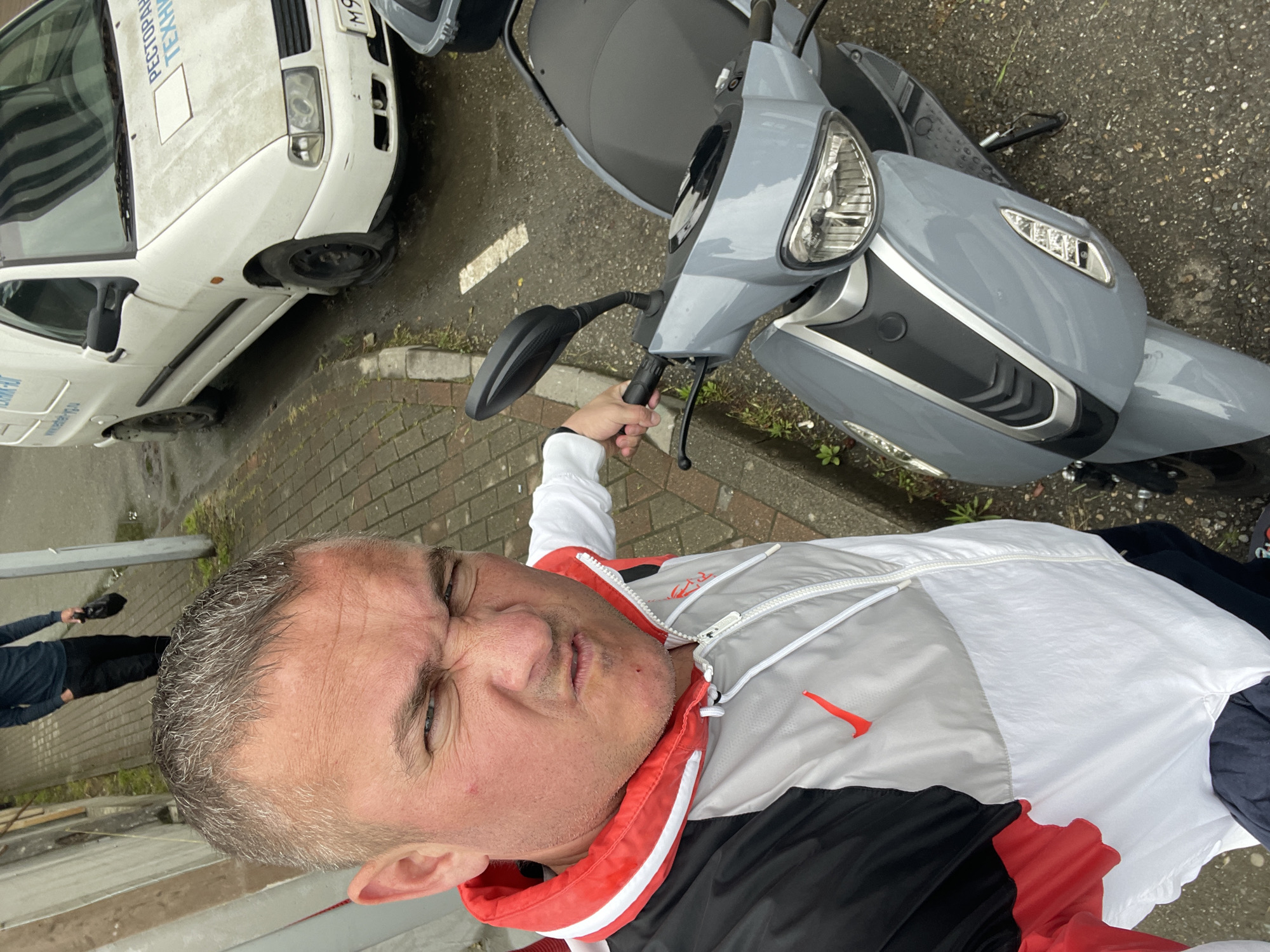 A selfie, taken from above, shows a man standing on what appears to be a street in Sochi, Russia. He is holding the handlebar of a gray scooter. A white van, labeled with "РЕСТОРАН ТЕХН", and a partial license plate beginning with "M9" is visible to the left. The man is wearing a white, gray, and black jacket with red accents and a visible brand logo. Another individual's feet are visible in the lower left of the frame. The scooter is parked on a paved surface.