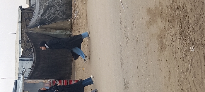 Two women are walking outdoors on a dirt road near what appears to be makeshift shelters or tents. Both women are wearing dark-colored garments and blue jeans. The terrain is somewhat barren, with the shelters seemingly constructed from tarps and other found materials. The sky is overcast, contributing to a sense of dreariness in the scene.