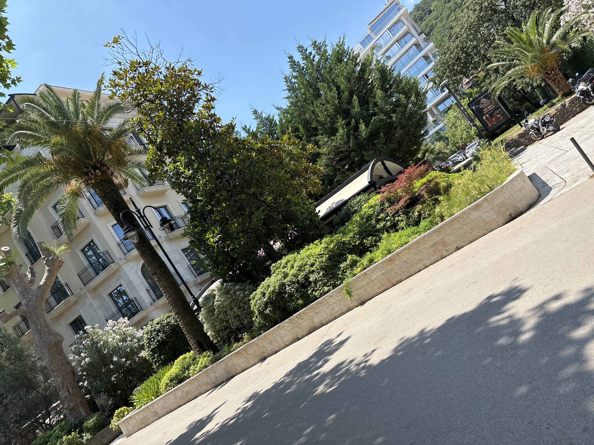 The image captures a street scene in Budva, Montenegro, oriented with the camera tilted. In the foreground, a paved roadway with tree shadows is visible, bordered by a stone retaining wall. Beyond the wall, a lush garden area features various plants, including dense green bushes, flowering shrubs, and a prominent palm tree. A multi-story building with a light-colored facade and balconies is situated behind the garden. Further in the background, another modern glass-fronted building is visible, suggesting a more developed urban area. Several motorcycles are parked along the side of the road. The sky is clear and blue, indicating a sunny day.