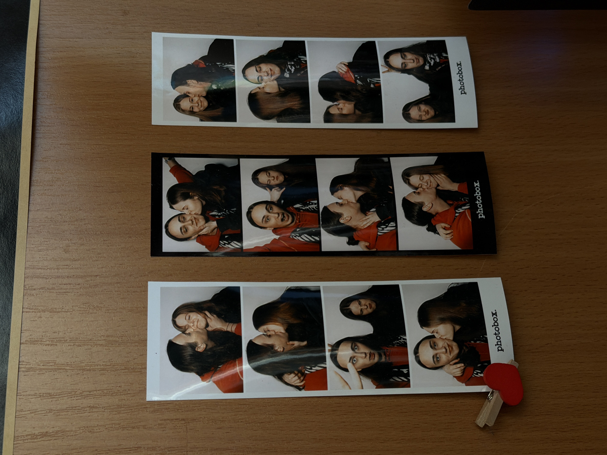 Three photobooth strips are on a wooden surface. The strips contain photos of two people, likely a couple, posing and kissing. The strips have "photobox." printed on them. One strip is secured with a small red heart-shaped clothespin clip. The location is not discernible from the image.