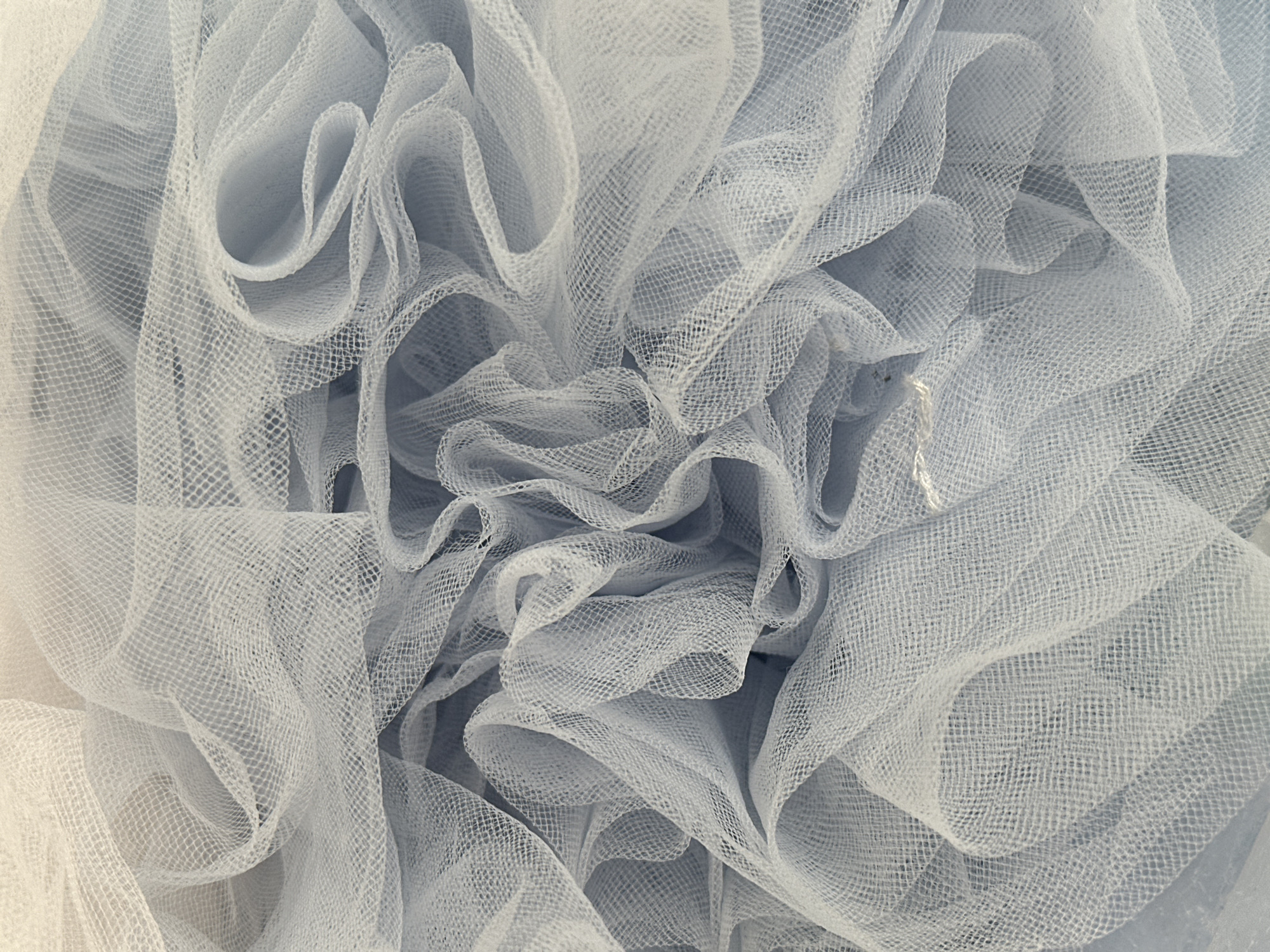 The image displays a dense, abstract arrangement of light blue-grey and off-white translucent mesh fabric. The material exhibits a fine, regular square-grid weave, characteristic of netting or tulle. It is extensively crumpled and folded, creating numerous undulating contours, overlapping layers, and deep recesses that dominate the entire frame. Variations in lighting accentuate the three-dimensional texture and the fabric's sheer quality, producing soft shadows within the folds. No other objects, persons, or distinct environmental features are discernible. The scene is located in Votkinsk, Russia.