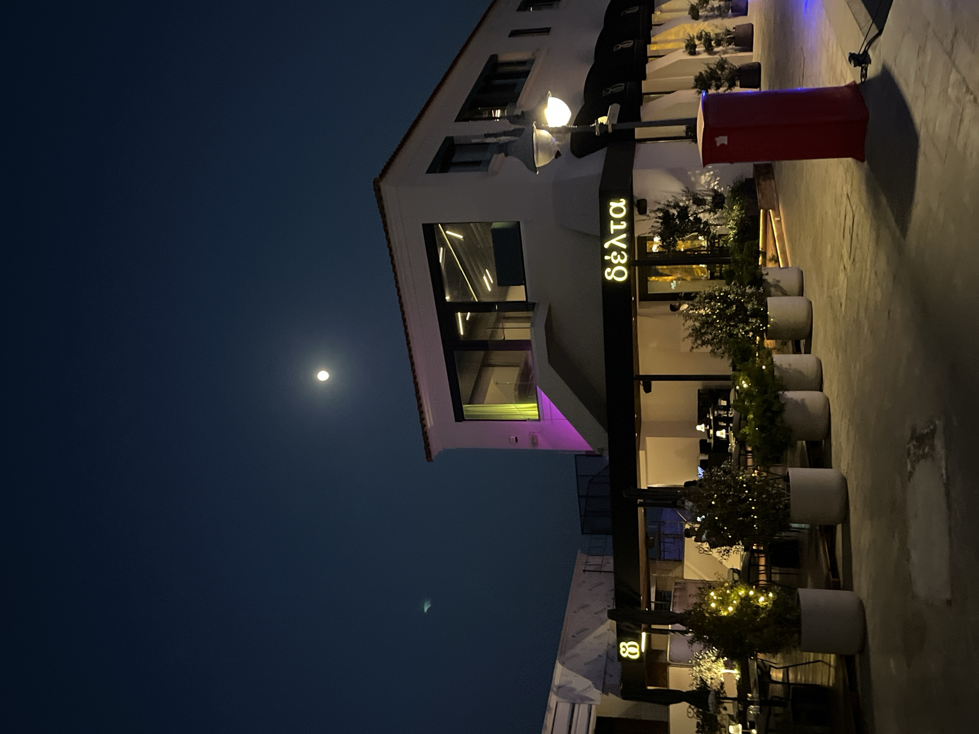 The image captures a nighttime view of a restaurant, likely named "Delta" in Limassol, Cyprus, indicated by the Greek lettering on the signage. The building is modern with large windows, and external lighting illuminates the facade with purple and yellow hues. In front of the restaurant, outdoor seating areas are visible with tables and chairs, surrounded by potted plants decorated with string lights. A prominent red post box stands on the paved area. The sky above is dark blue, featuring a bright full moon. In the distance, there appears to be another building with a logo that resembles the number 8.