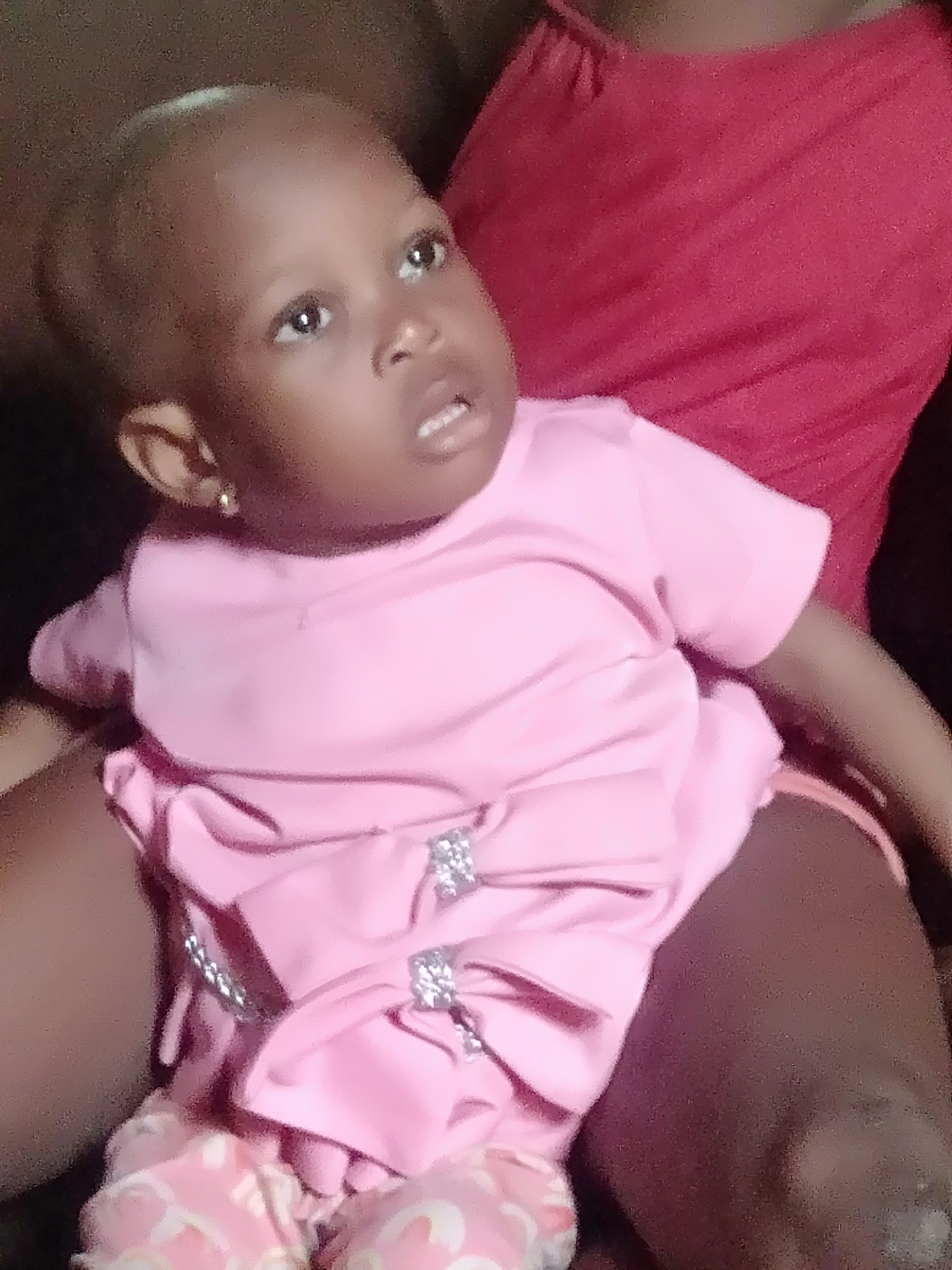 A close-up shot depicts a young child, presumably a baby or toddler, dressed in a pink outfit featuring decorative bows with small, glittering embellishments. The child sits on the lap of an individual wearing a red top. The child is looking up and to the side with a slightly open mouth, revealing a few teeth. A small, gold earring is visible on the child's ear. The location is Unknown, Unknown.