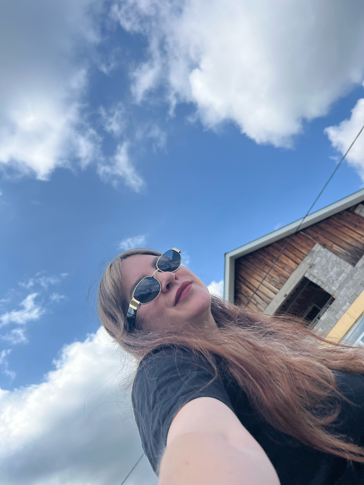 The image is a low-angle shot of a woman in Noviy, Russia, looking up at the sky. She is wearing sunglasses and a black t-shirt. Her hair is long and reddish-brown, and it falls over her shoulders. In the background, there is a partially constructed building with wooden siding. The sky is blue with scattered white clouds. The overall impression is one of a casual, outdoor moment.