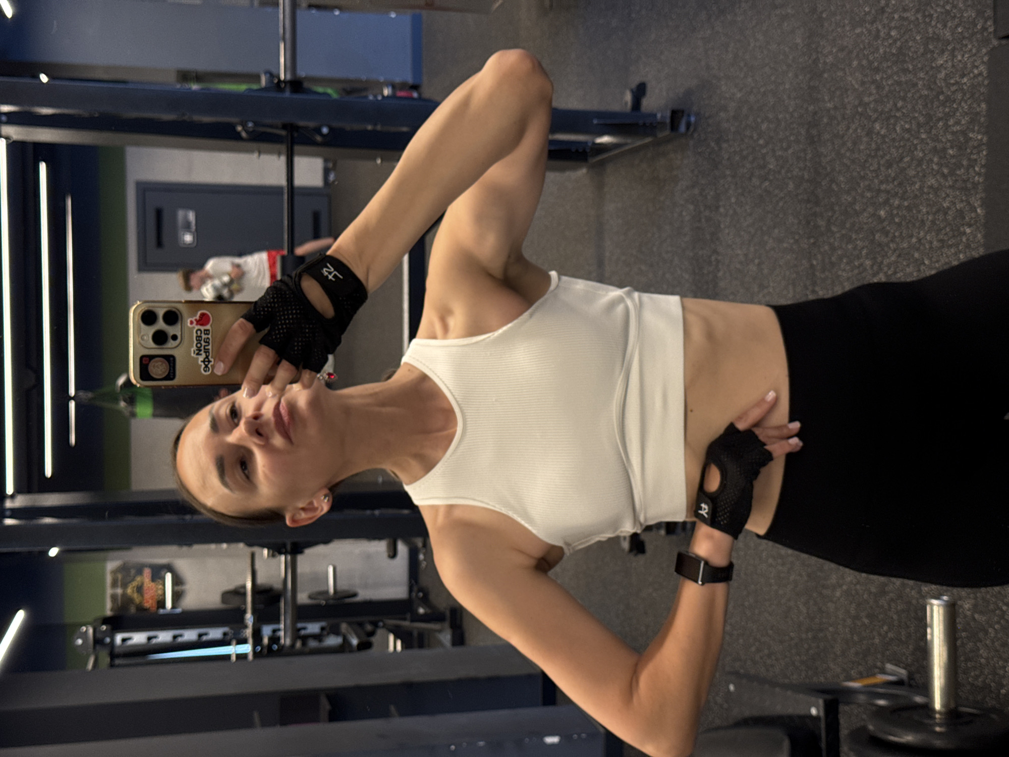 A woman is taking a mirror selfie in a gym. She is wearing a white ribbed tank top, black leggings, and black workout gloves. Her hair is pulled back, and she is wearing earrings and a watch. She is holding a smartphone in her left hand, which is angled to capture her reflection. In the background, equipment for weightlifting is visible, including barbells and weight plates. The gym floor is dark grey and textured. A reflection of another person, wearing a white shirt and shorts, can be seen in the mirror in the distance. The lighting suggests it is daytime or that the gym is well-lit. The woman's expression is neutral, and she appears focused on taking the photo. The phone case has some text that appears to be in Russian.