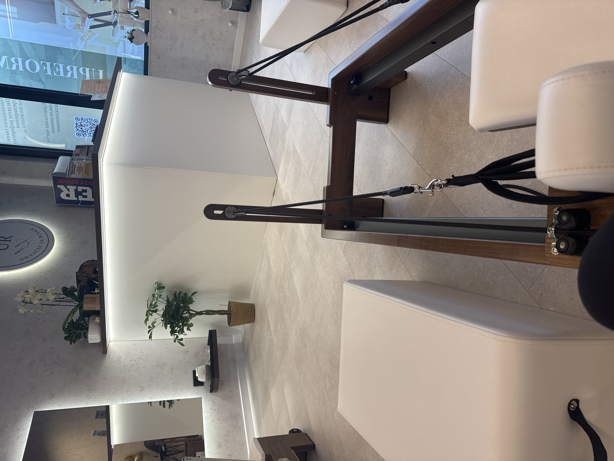 Here is a detailed description of the image:
**Scene:**
The image shows the interior of what appears to be a Pilates or fitness studio. The main focus is on a Pilates reformer machine, which is made of dark wood and has white padded surfaces. Black cables and pulleys are visible, connecting different parts of the machine.
**Objects:**
* **Pilates Reformer:** The central object is the Pilates reformer machine, designed for exercise and rehabilitation.
* **White padded benches/platforms:** In addition to the reformer, there are white padded benches or platforms around, likely for other exercises or waiting areas.
* **Plants:** A small potted plant is placed on a shelf along the wall, adding a touch of greenery to the space.
* **Mirrors:** A mirror is visible near the floor, suggesting this is a place for physical activity where one would want to see their reflection.
* **Signage:** A sign in the background has the words "ПИЛАТЕС".
* **Light fixtures:** The studio has a modern lighting design with integrated lighting along the walls and shelves.
**Environment:**
The studio has a clean, modern aesthetic with light-colored walls and flooring. The space is well-lit and appears to be well-maintained.
**Possible Location:**
Based on the text from signage, "ПИЛАТЕС ЗА ВСЕКИ" and "RHCOKOKIAGGLBGHH", the image may be from a Pilates studio in Sofia, Bulgaria. This is only a possibility.