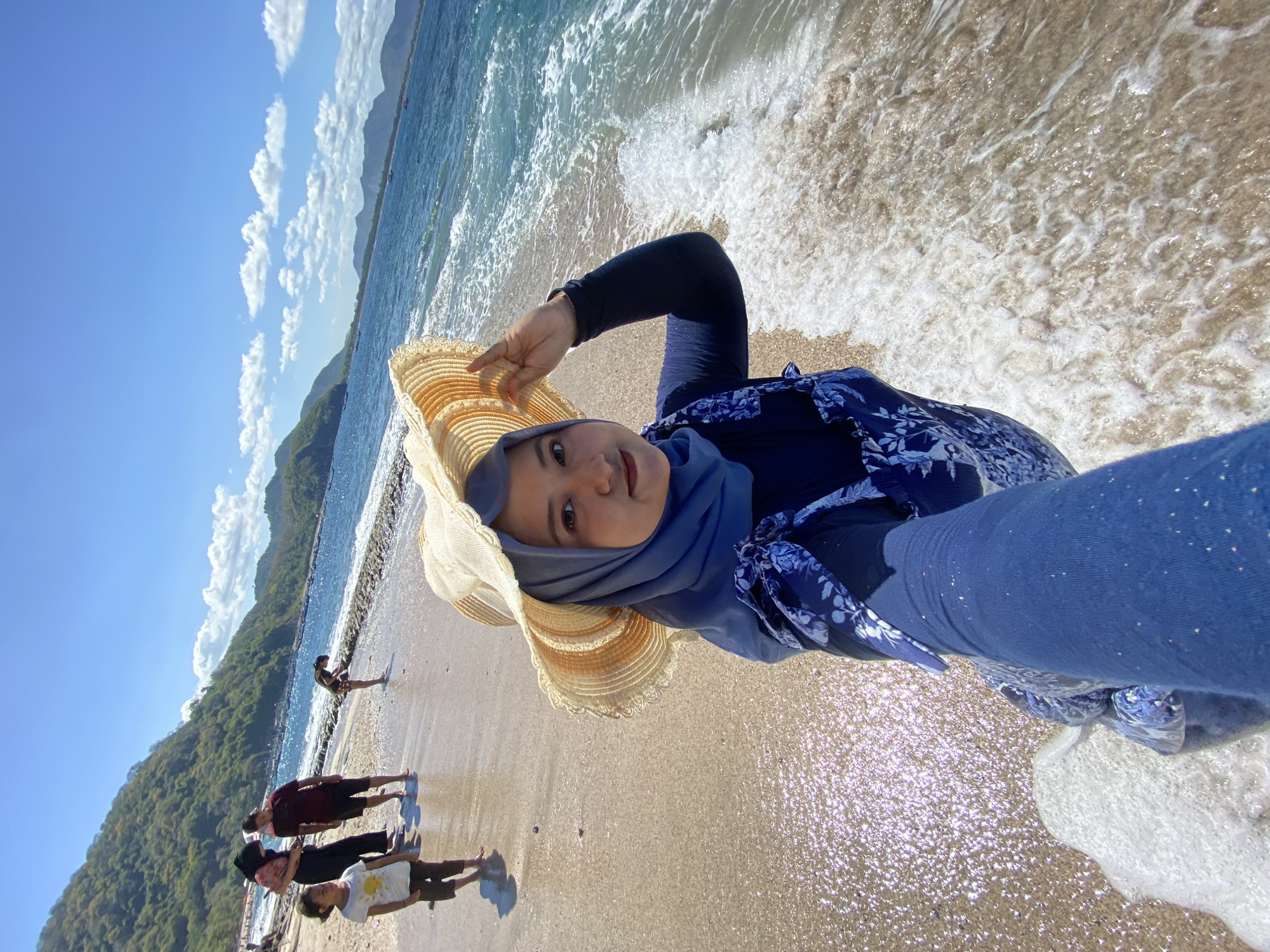 A young woman wearing a sun hat and head covering stands on a beach, possibly in Unknown, Indonesia, as suggested by the setting. She is positioned at an angle facing the camera. Behind her, the shoreline is visible, with several other people standing and walking along the beach. The ocean waves are lapping at the shore. A densely forested hill or mountain is present in the background under a clear, sunny sky with some clouds.