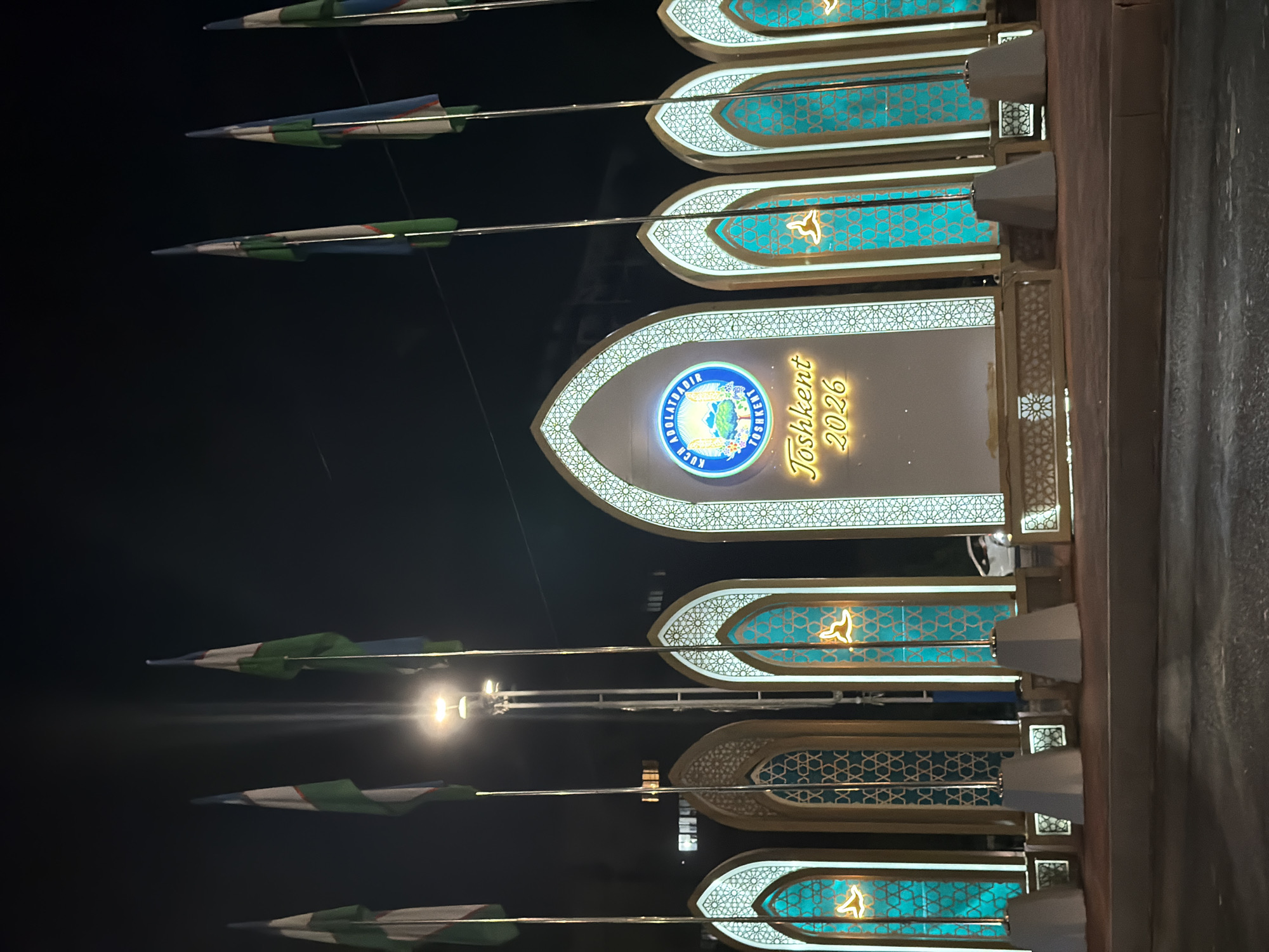 The image shows an outdoor nighttime scene in Tashkent, Uzbekistan, featuring a large, illuminated monument. The monument is flanked by multiple flags on poles, which appear to be the national flag of Uzbekistan. The monument itself is decorated with Islamic architectural motifs, including arched doorways and intricate geometric patterns. In the center of the monument, a circular emblem is visible, along with the text "Toshkent 2026" illuminated in a golden script. The overall impression is of a festive or commemorative display, possibly related to an upcoming event in Tashkent. The background is dark, suggesting it is late at night, with some faint building lights visible in the distance.