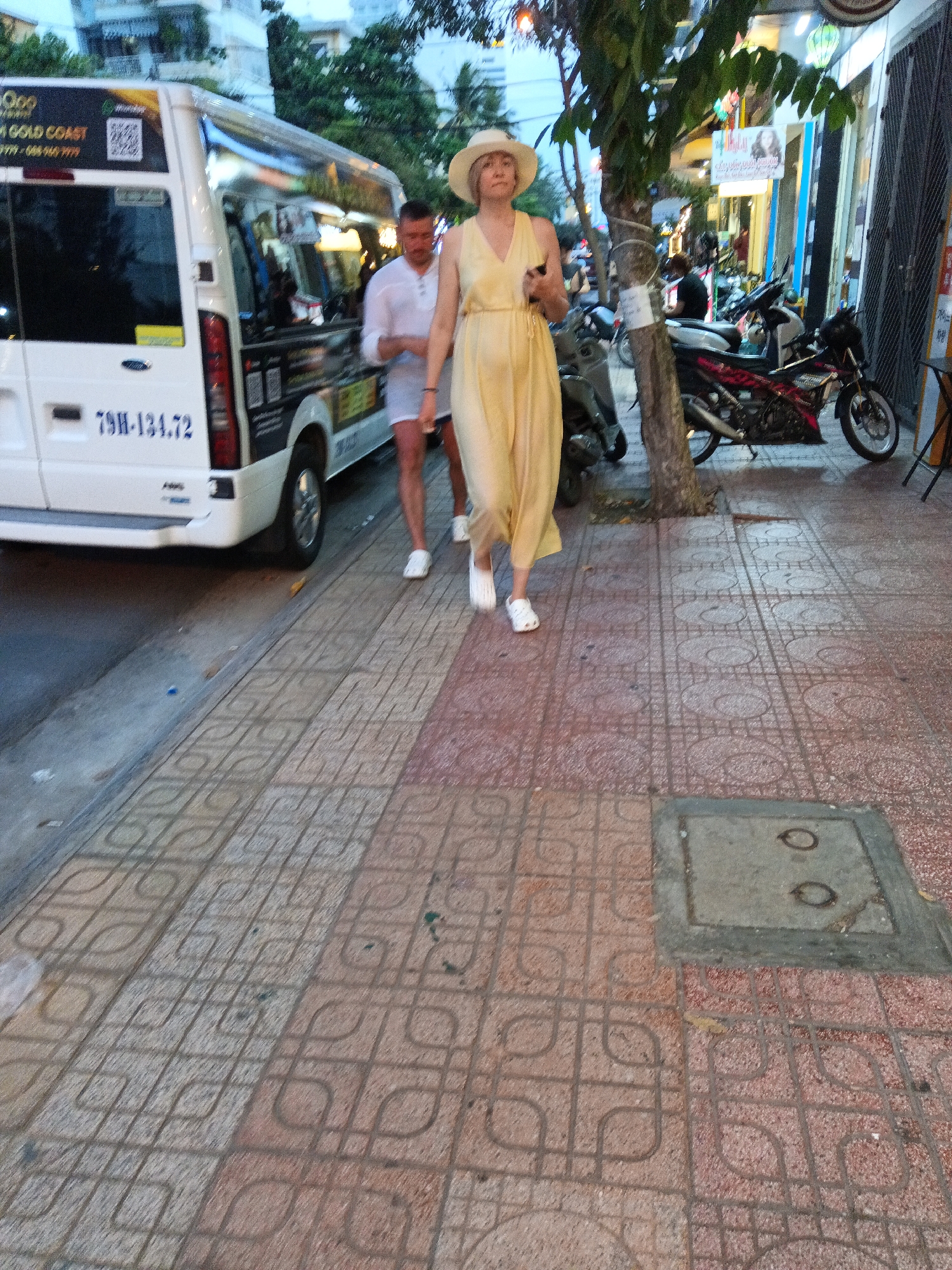 In Nha Trang, Vietnam, a white van with the license plate "79H-134.72" is parked on the side of a street. A man in a white shirt and shorts stands next to the van, and a woman in a light yellow dress and a white hat walks on the sidewalk. The sidewalk is paved with red and gray tiles. There are trees and buildings in the background. The scene appears to be in the evening.