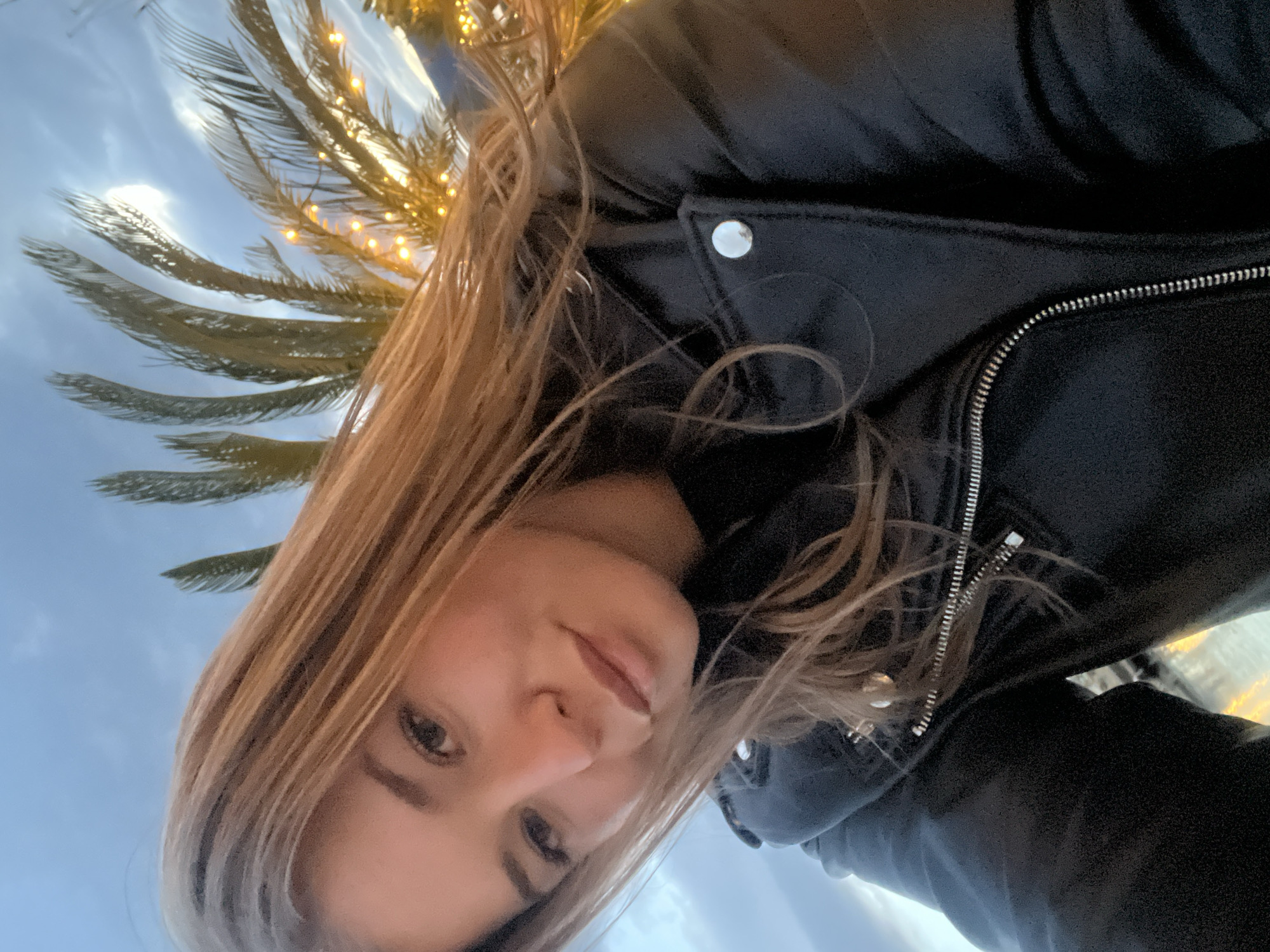 A selfie taken from a low angle features a young woman with light brown hair, wearing a black leather jacket with a silver zipper and a button visible on the upper part of the jacket. Behind her is a palm tree decorated with small, warm-toned lights. The sky, visible in the background, is a light blue with some cloud cover. Considering the context of the city of Tivat, Montenegro, it's plausible the palm tree is a decorative element along the marina or town center.