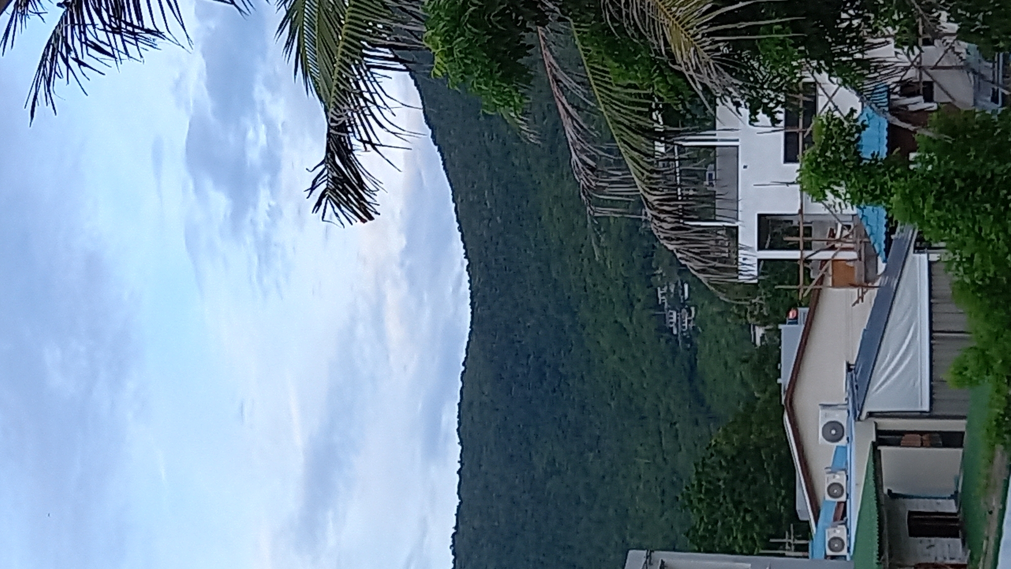 The image depicts a tropical landscape in the Ko Pha-ngan District of Thailand. In the foreground, several low-rise buildings with various rooflines are visible, including structures with white siding and tiled roofs, as well as what appears to be a construction site with scaffolding. Air conditioning units are mounted on one of the building's roofs. Dominating the middle ground is a lush, densely vegetated hillside, characteristic of the region's natural environment. In the distance, nestled within the hillside, are several small buildings, possibly residences. The sky above is partly cloudy, with a soft, diffused light suggesting either early morning or late afternoon. The fronds of a palm tree are prominently featured in the right side of the frame, adding to the tropical ambiance. The scene is static, with no visible people or animals.