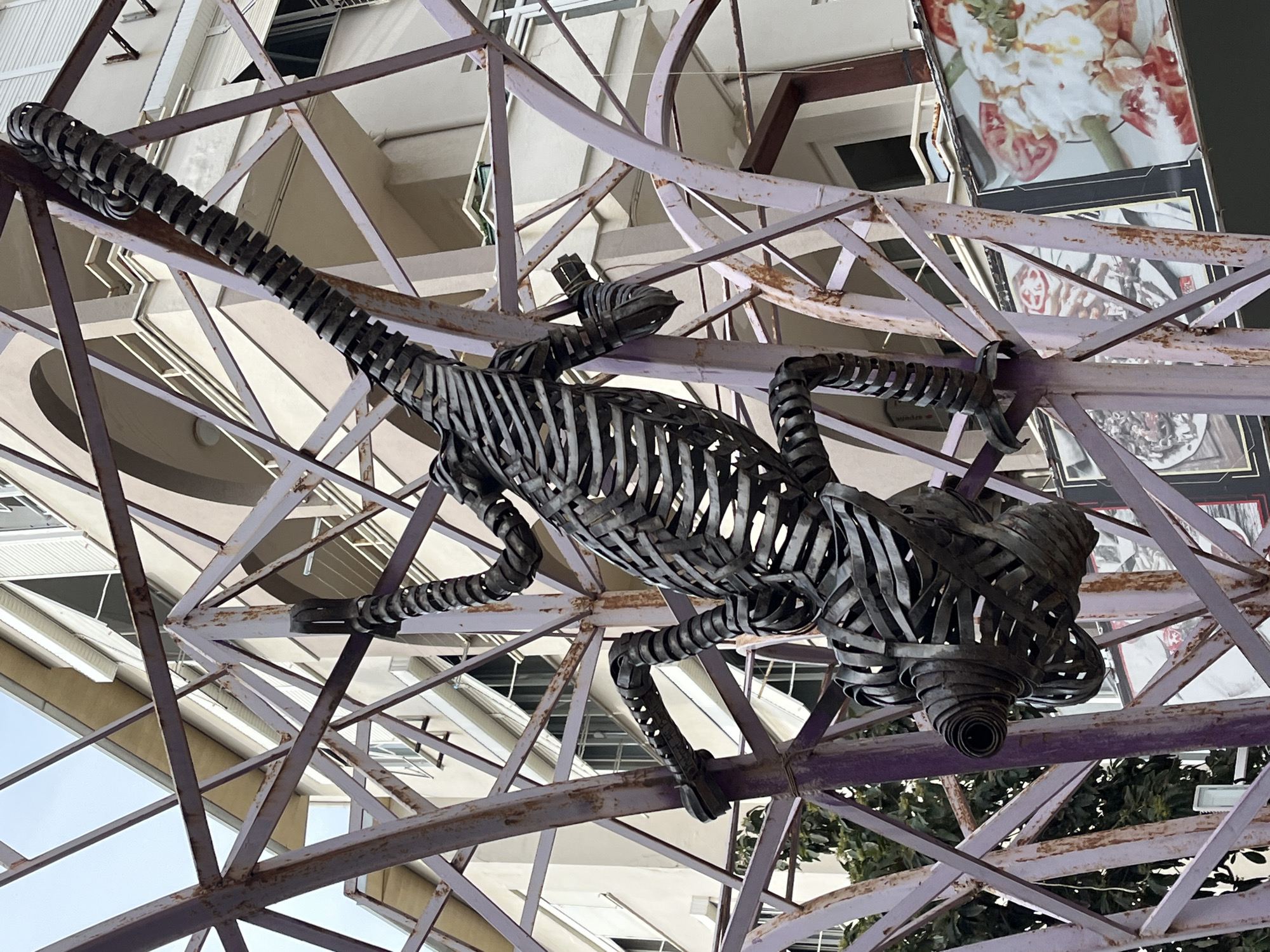 The image shows a metal sculpture of a reptile, possibly a lizard or crocodile, suspended from a purple metal framework. The sculpture is made of metal strips and bands. The metal framework consists of intersecting beams and is visibly rusted in places. The background includes architectural elements of buildings, such as windows and facades. The presence of food-related advertisements, like the pizza advertisement in the background, indicates the location is near a restaurant or food establishment. Given the urban environment, the sculpture appears to be a public art installation. The location is Alanya, Turkey.
