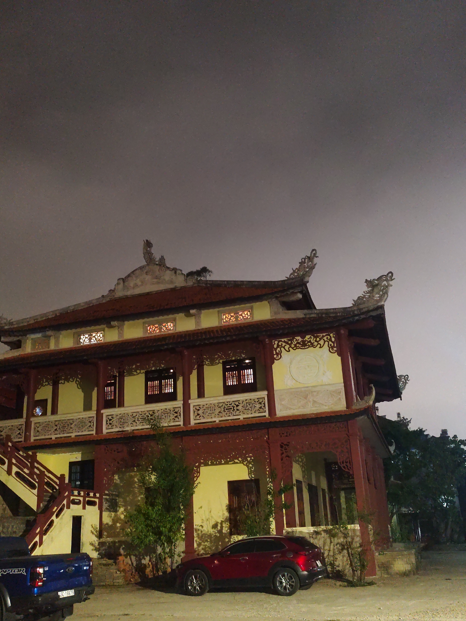 The image shows a two-story traditional Vietnamese building with yellow walls, a red roof, and intricate carvings, likely a temple or historical site. Two vehicles, a blue Ford Raptor pickup truck and a red Mazda SUV, are parked in front of the building. The sky is overcast and dark, suggesting it is nighttime. The building is located in Da Lat, Vietnam.