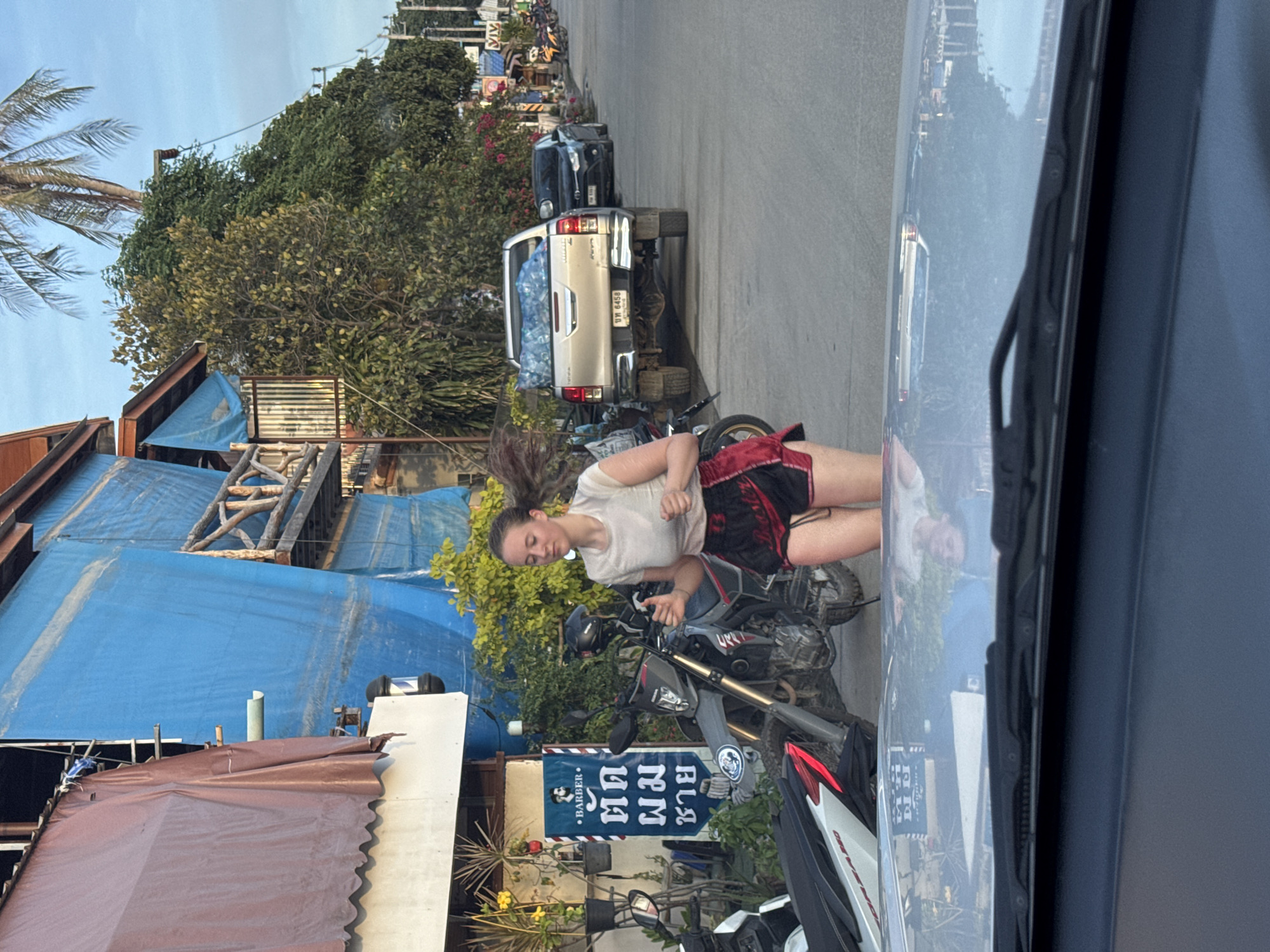 A young Caucasian woman with blonde hair pulled back runs on a paved street in front of a building covered with blue tarp. She wears a white t-shirt and black shorts with red trim. Behind her are two motorcycles and a barber shop sign with Thai writing. A silver pickup truck is parked further down the street, followed by other vehicles and shops. The scene is outdoors, with trees and a palm tree visible. The sky is partly cloudy. The image appears to be taken from inside a vehicle.