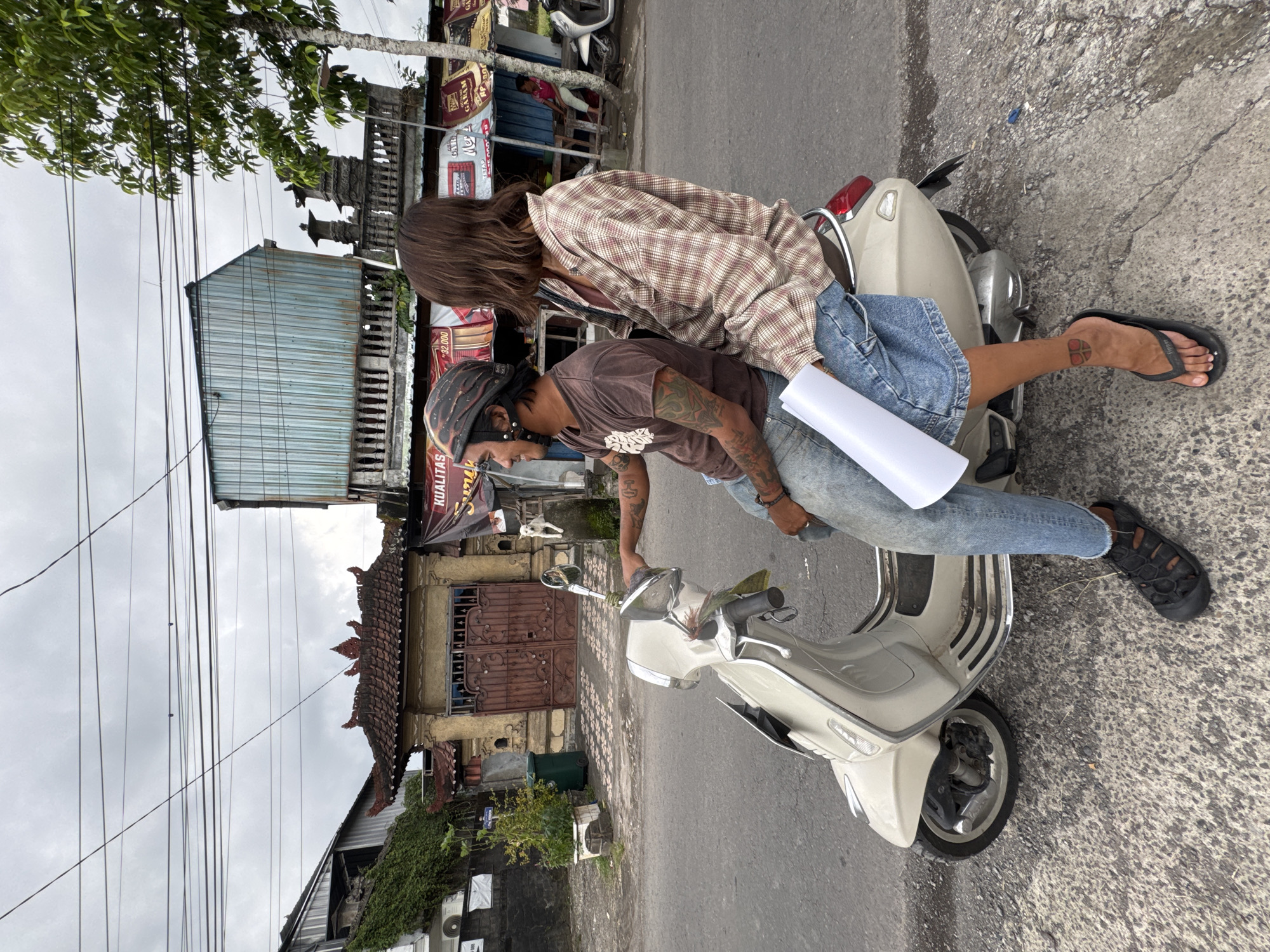 In a city street scene in Kediri, Indonesia, two individuals are interacting near a white Vespa scooter. The scooter is parked on cracked asphalt. One person is seated on the scooter, wearing a brown shirt, light blue jeans, a helmet, and black shoes. This individual is partially covered in tattoos on their arms. The second person is leaning over the seated person, wearing a tan plaid shirt and blue denim shorts. Their leg, adorned with a tattoo, is also visible. The street is bordered by various buildings, some with traditional Indonesian architectural details. A white cylinder of paper is visible between the two individuals. Overhead power lines run parallel above the street, and lush greenery is present in the scene.