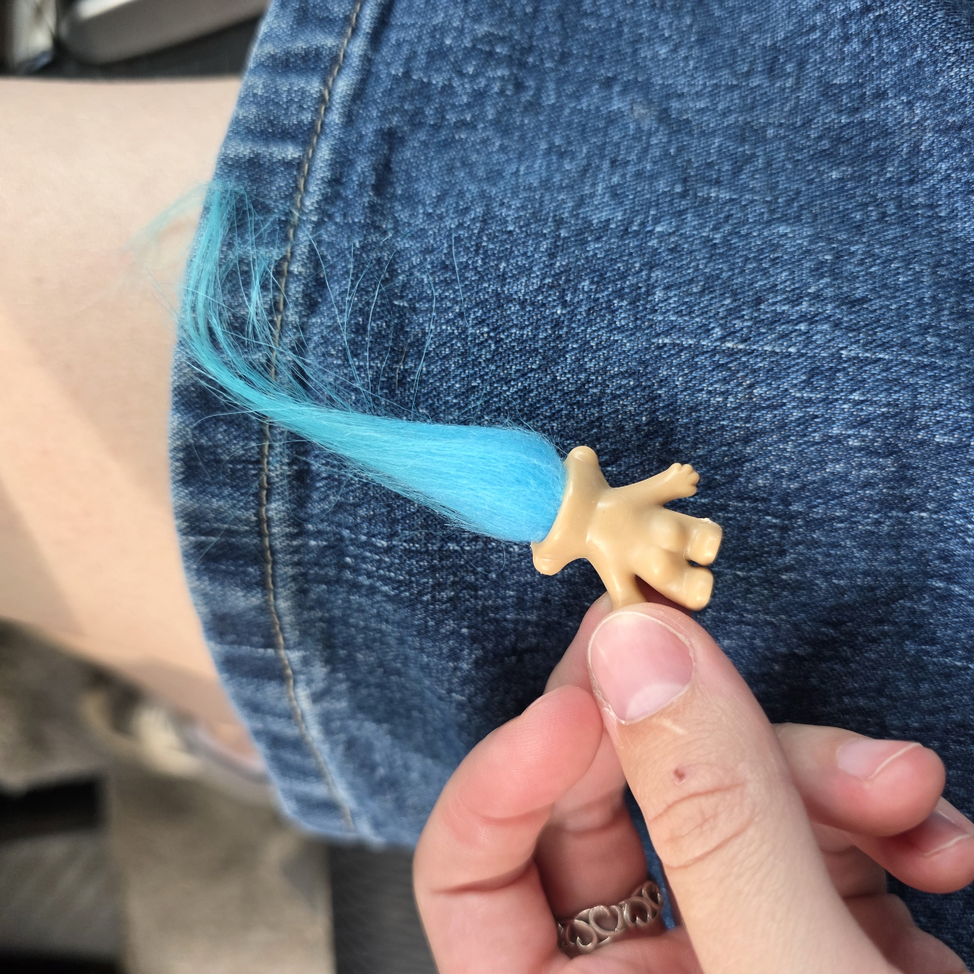 A close-up shot focuses on a hand holding a small, light-tan plastic doll arm with bright blue hair sticking out of the end. The doll's arm appears to be that of a troll doll. The hand, belonging to a person, is wearing a silver ring with a heart-shaped cutout pattern on the ring finger. The background consists of a textured blue denim fabric, likely jeans, and a hint of skin on the left side, possibly an arm or leg. The lighting suggests it is daytime, with natural light illuminating the scene. The image has a casual, everyday feel. No text is visible in the image.