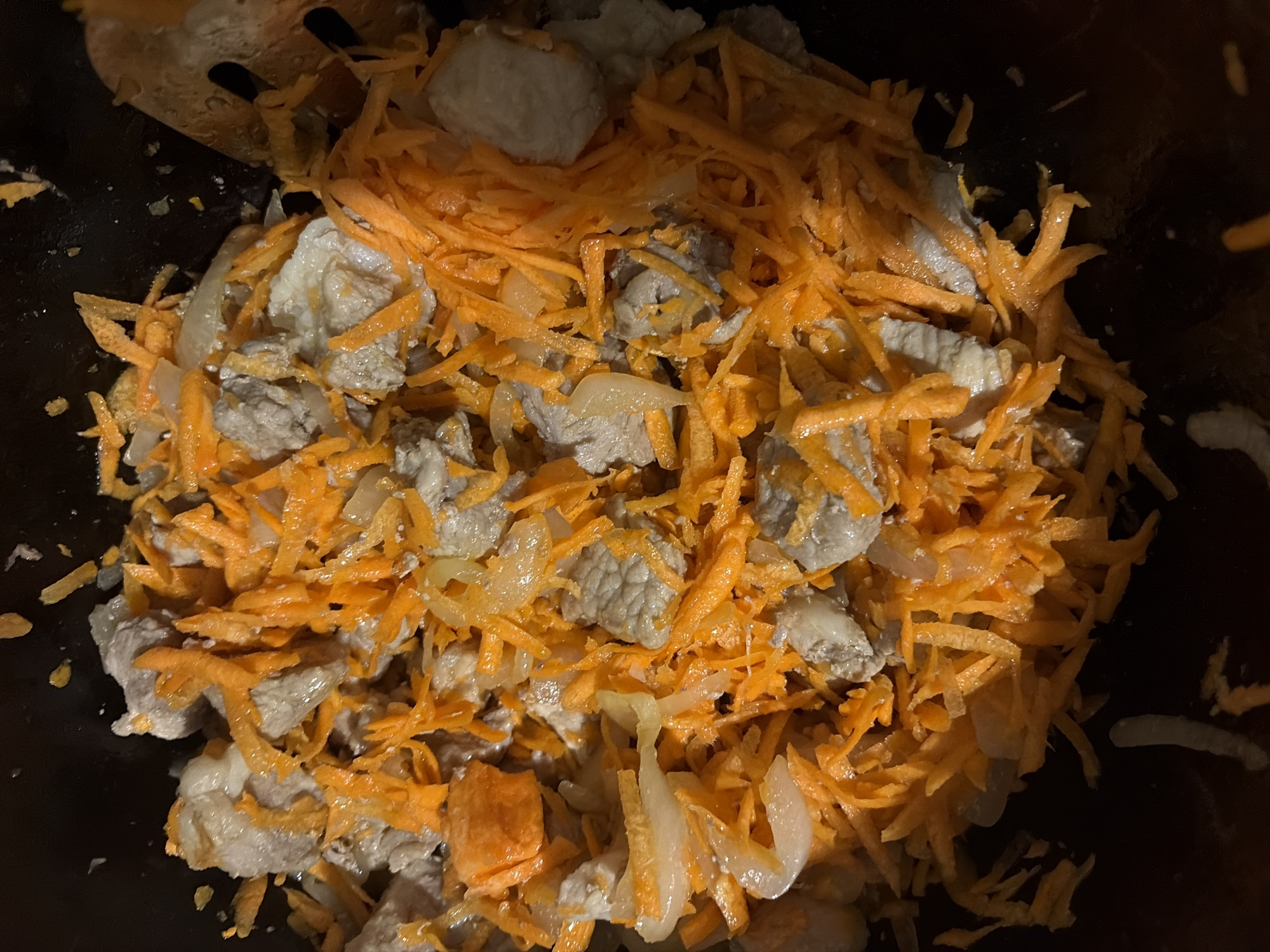 An overhead close-up shot shows a dark pot filled with a mixture of shredded carrots, sliced onions, and cubed pieces of meat. The ingredients are partially cooked, with the meat appearing gray and the carrots and onions translucent. Some of the meat has a slightly browned exterior. The ingredients are unevenly distributed, creating a textured surface. The pot's dark interior is visible around the edges, suggesting it's a cooking vessel. The lighting is dim, casting shadows and highlighting the sheen of the ingredients. The scene depicts food preparation, likely in a kitchen setting in Tambov, Russia.