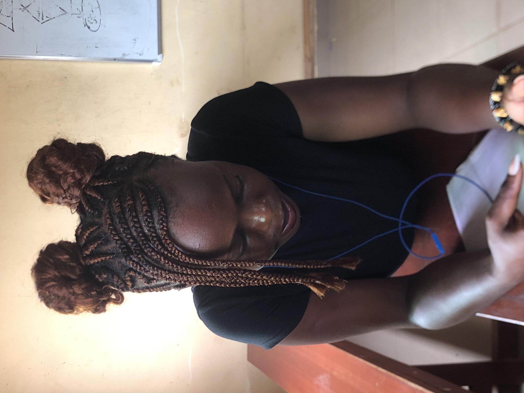 The image depicts a dark-skinned woman with braided hair styled in two buns. She is wearing a black shirt and appears to be looking down at a piece of paper she holds in her hands. Blue wired earphones are visible near her neck. She is sitting at a wooden desk or table. A whiteboard with writing on it is partially visible in the background. The location is Nsugbe, Nigeria.