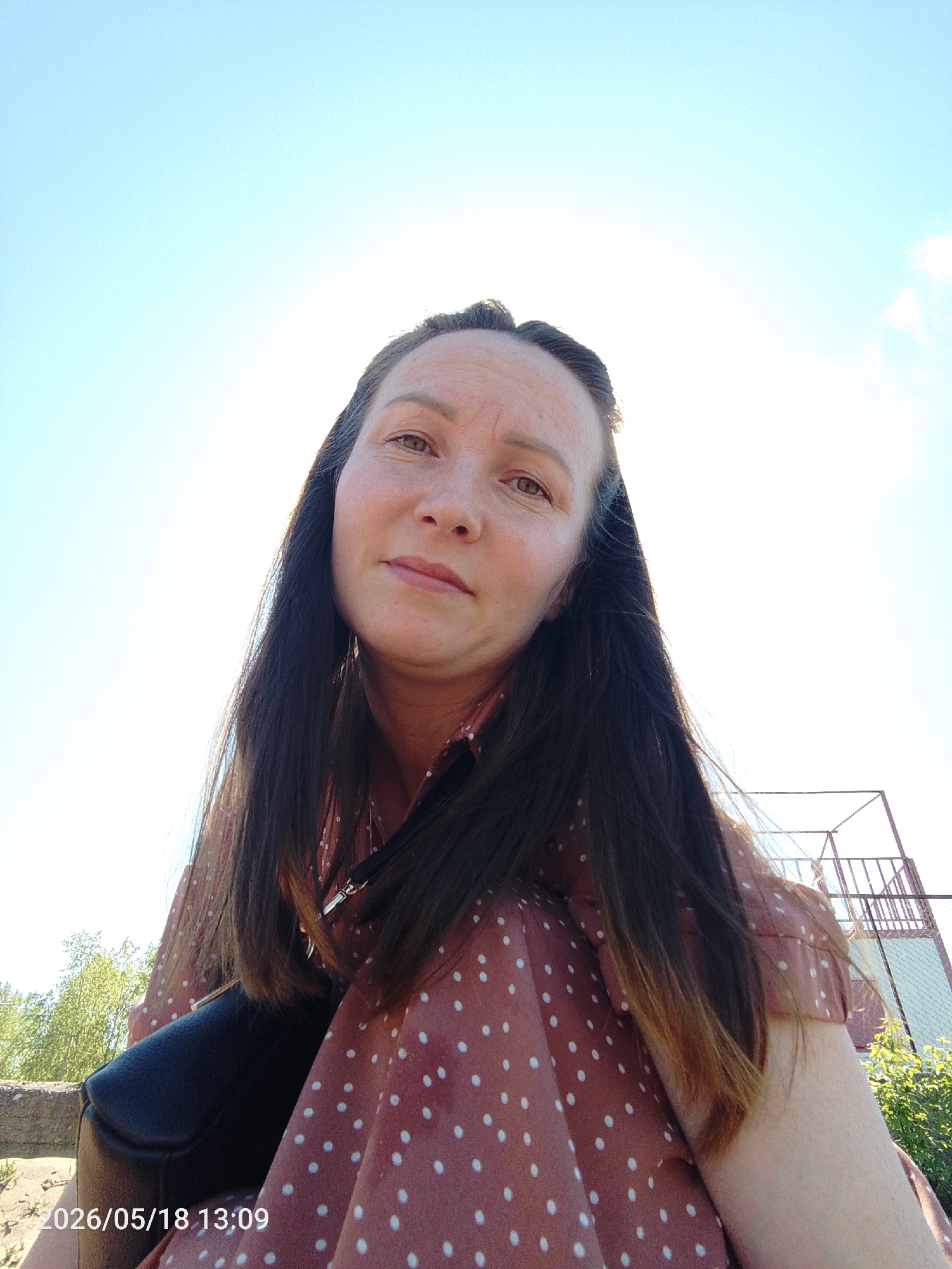 This outdoor, low-angle selfie captures a woman looking directly at the camera under a bright, clear blue sky. She has long, dark brown hair, a neutral to subtly pleasant expression, and light brown eyes. She is wearing a reddish-brown or terracotta-colored top adorned with white polka dots. A black handbag is visible on her lap or beside her.
The background shows sparse green trees or foliage on the left and a metal structure, possibly a fence or part of an outdoor installation, on the right. The bright sunlight creates significant glare in the upper right portion of the sky, indicating a very sunny day. The timestamp "2026/05/18 13:09" is visible in the bottom left corner, marking the date and time as early afternoon. The overall scene suggests an outdoor setting in Chaikovsky, Russia, on a clear and sunny day.