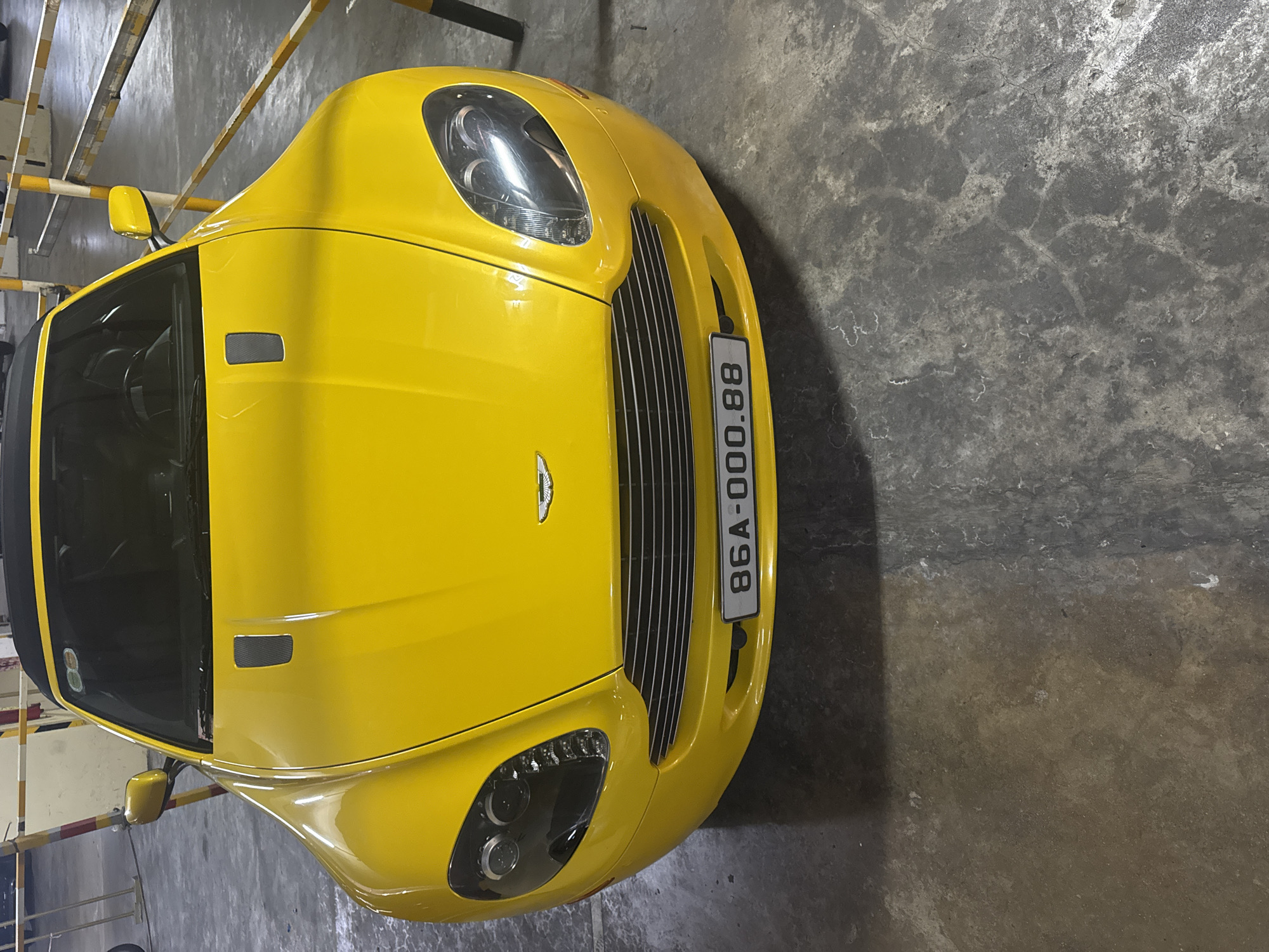 A bright yellow Aston Martin Vantage is parked in a dimly lit parking garage. The car's front is facing the camera, showcasing its sleek design, including its distinctive grille, headlights, and a license plate reading "86A-000.88". The car is positioned on a concrete floor that appears weathered and slightly wet in places. Yellow and white striped barriers are visible in the background, suggesting this is a designated parking area. The overall lighting is low, with artificial lights casting shadows. No people are present in the image. The location is identified as Nha Trang Ward, Vietnam.